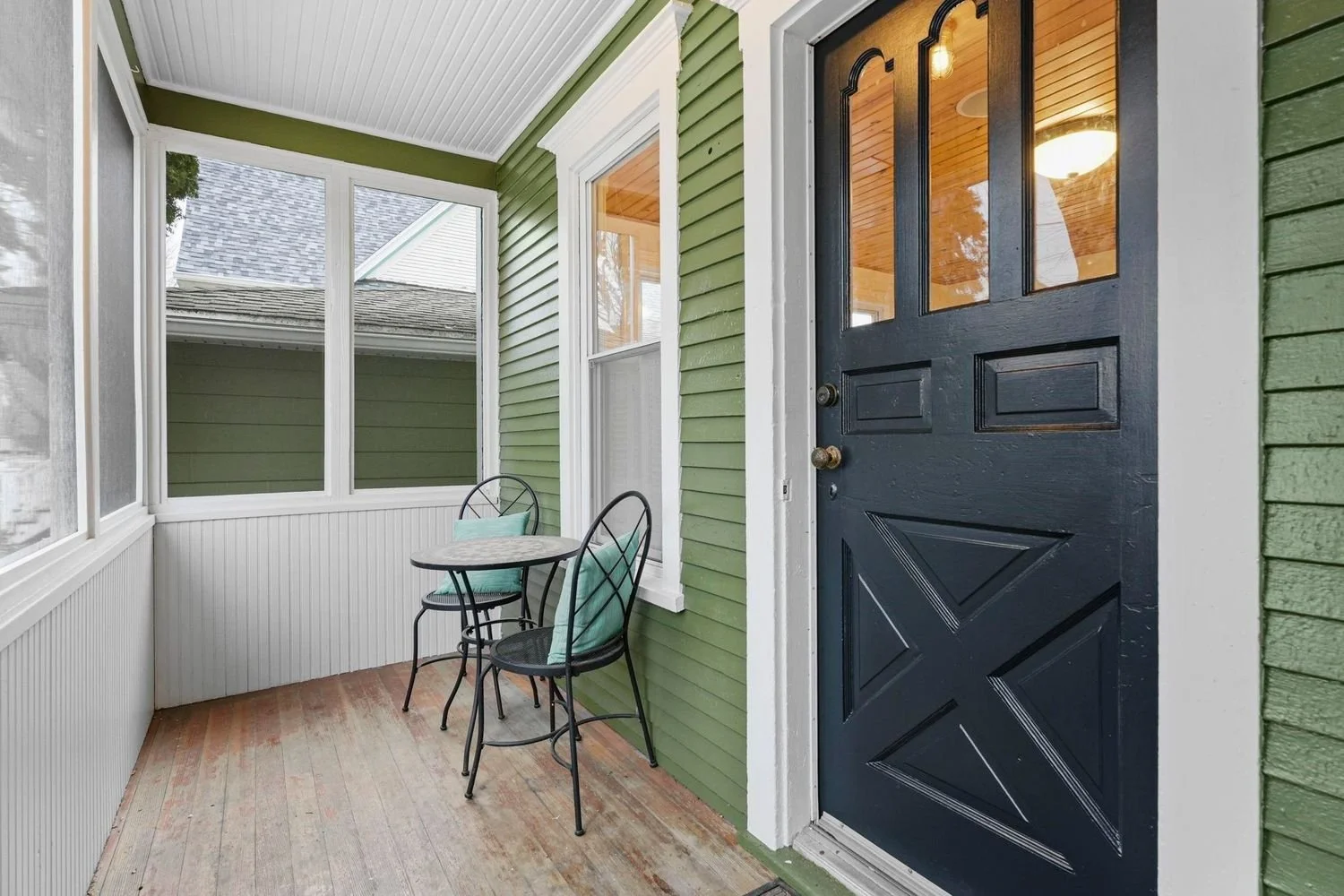 A small porch with a black door, green exterior walls, and a white ceiling. There is a small round table with two metal chairs, each with a turquoise cushion. The porch has large windows with white trim and a wooden floor.
