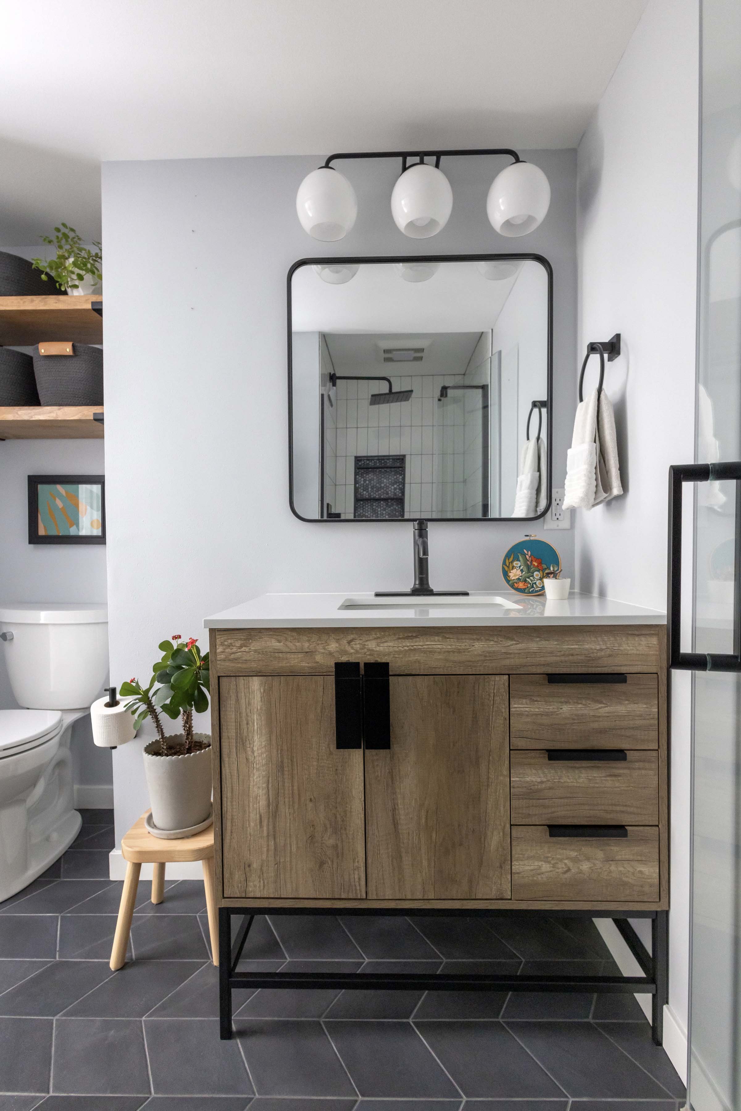 Modern bathroom with white walls and black tiled floor, featuring a wooden vanity with black accents, a white countertop with an integrated sink, and a large mirror reflecting a shower with square tiles and a black shower head. The bathroom has a minimalist aesthetic with small plants and decorative items.