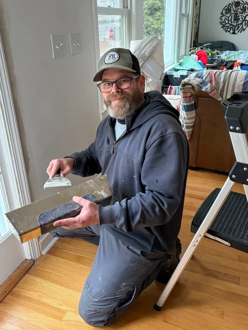 A man wearing glasses and a baseball cap kneeling in front of a sunlit window, holding a drywall mudding tool and a sponge, with a ladder nearby. There are household items and furniture in the background.