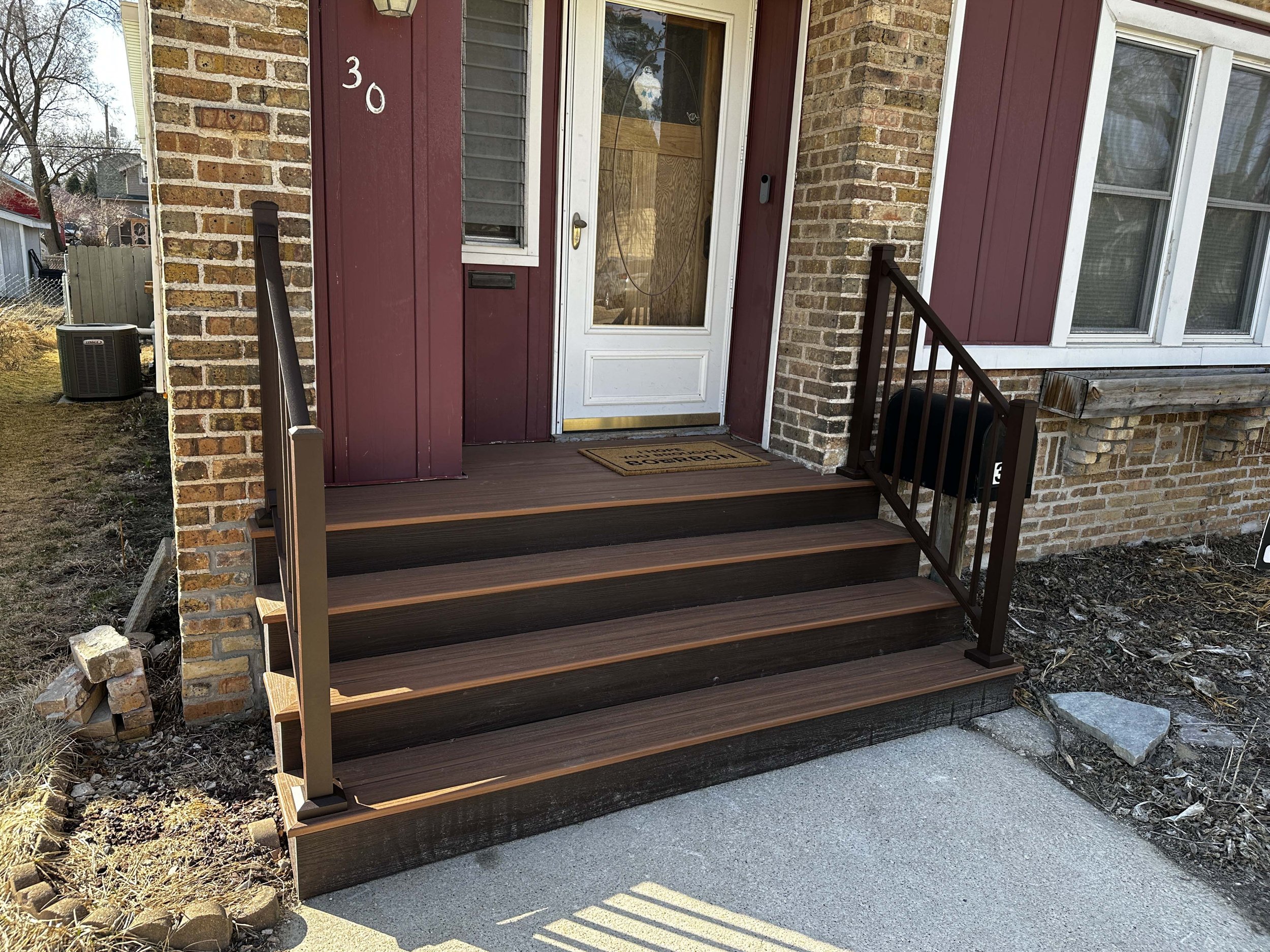 Front porch of a house with a new wooden staircase with black metal railings leading up to a white door with a glass window. The house has red and tan brick exterior walls, and a small window is visible to the right of the door. There is an outdoor air conditioning unit on the left and some landscaping with rocks and soil.