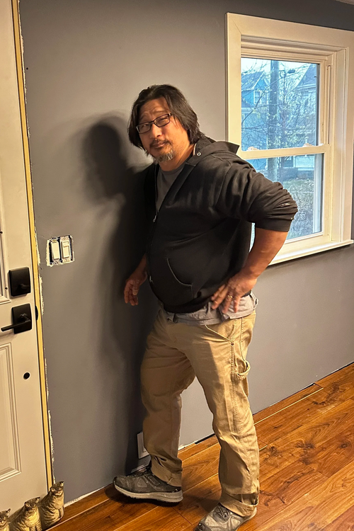 A man with shoulder-length dark hair, glasses, and facial hair stands next to a gray wall, with a window showing outdoor trees and houses in the background. He is wearing a black hoodie, beige cargo pants, and sneakers, with one hand on his hip.