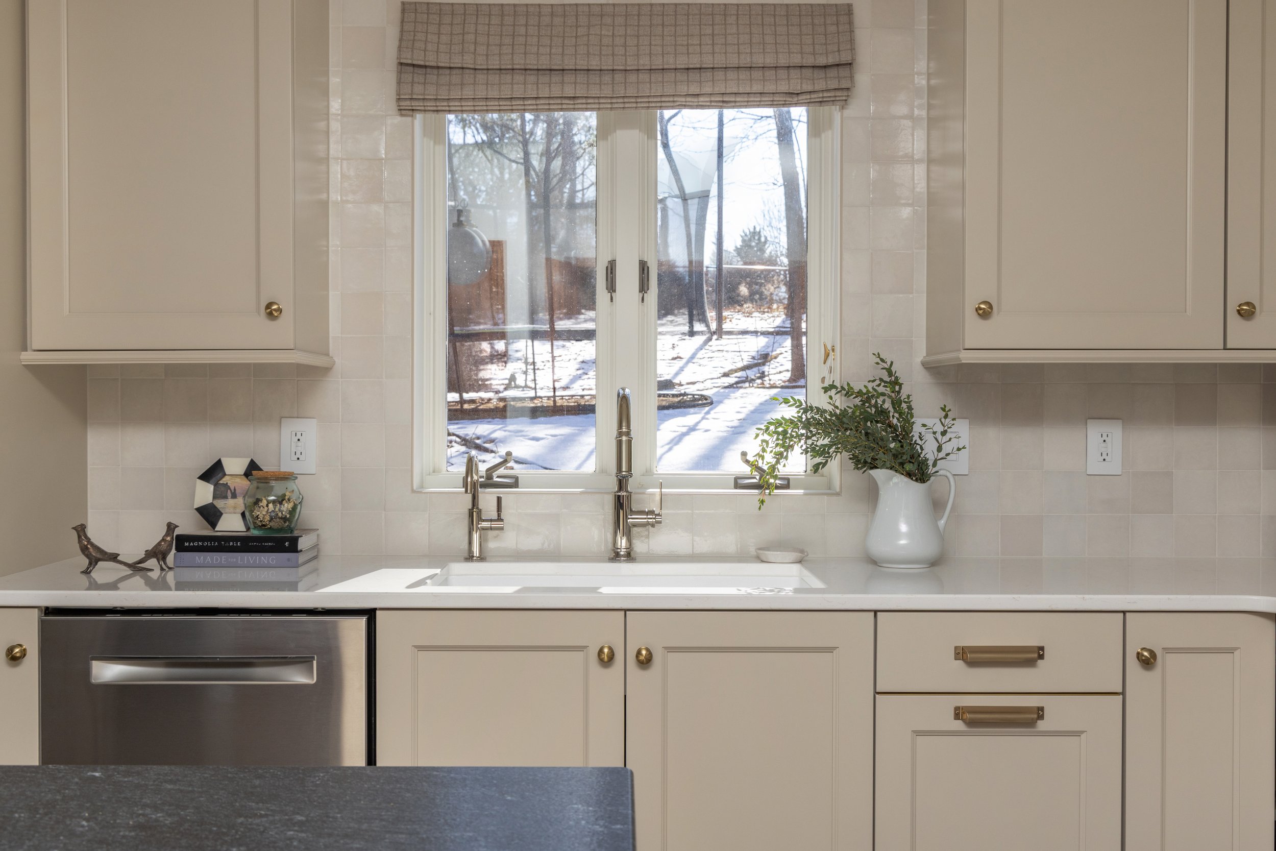 A kitchen with a window showing a snowy outdoor scene, white cabinets, a white countertop, and various decorative items including a plant in a white pitcher and books.