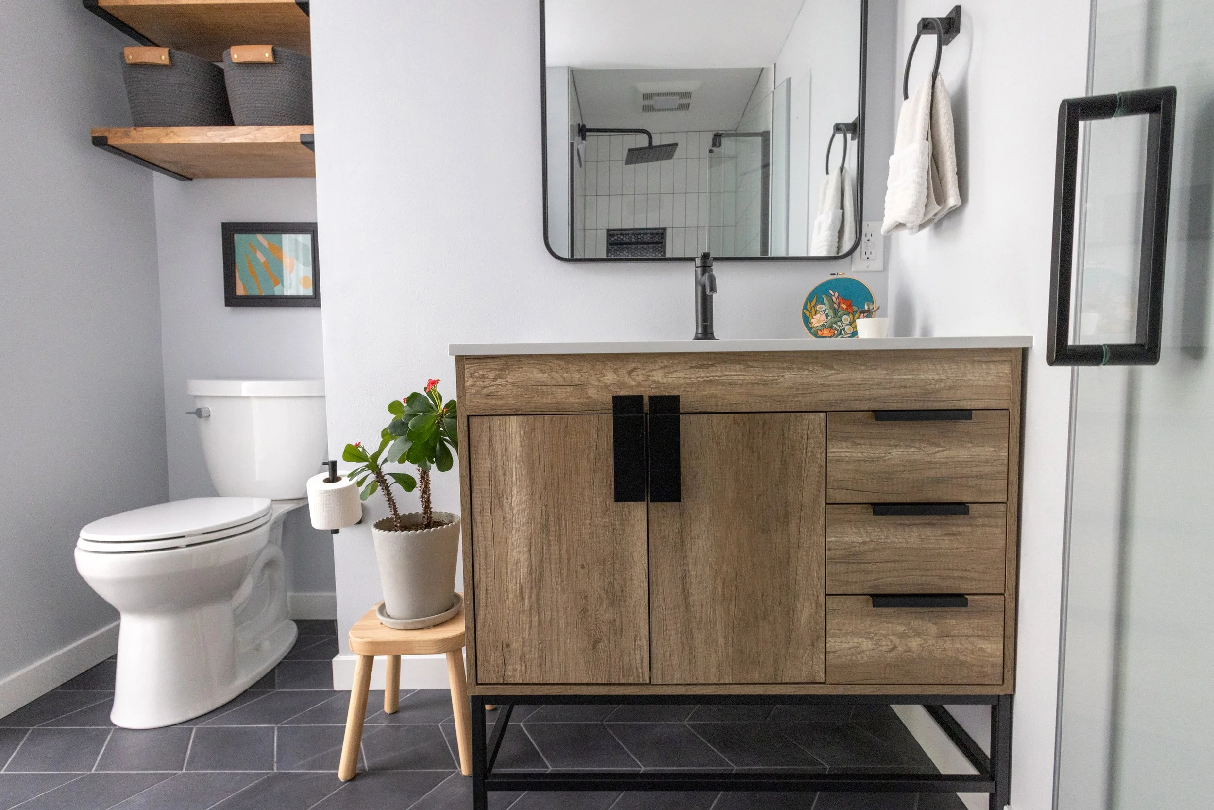 Modern bathroom with a toilet, a wooden cabinet with black handles, a large mirror, a black faucet, and a shower area with a glass door. The bathroom is decorated with a small plant, a colorful plate, and towels.