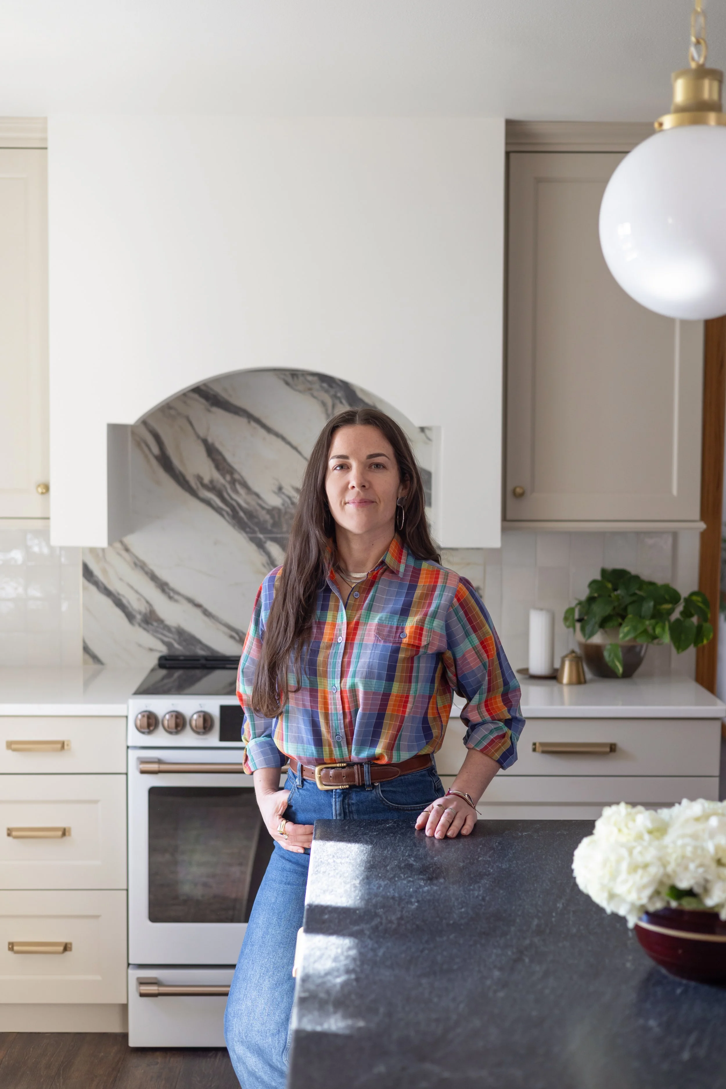 A woman with long dark hair in a plaid shirt and jeans standing in a modern kitchen, resting her hand on a black countertop with a bowl of white flowers in the foreground, cream-colored cabinets, an oven with copper knobs, a marble backsplash, and a white hanging light fixture.