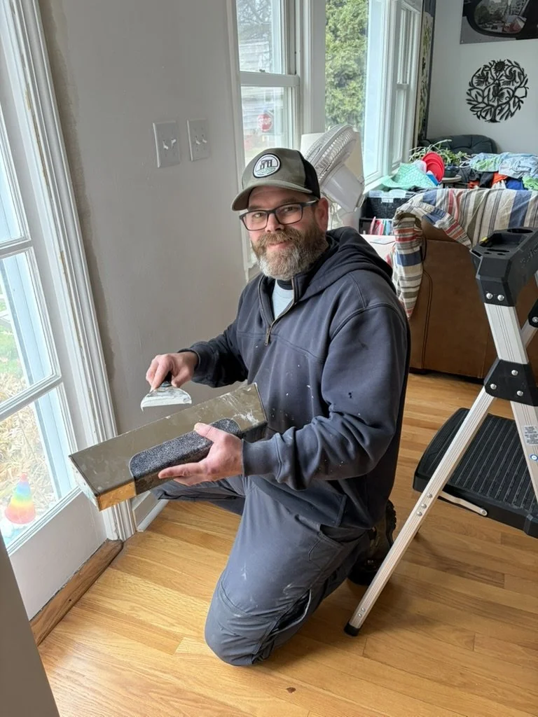 A man with a beard and glasses kneeling on a hardwood floor, holding a squeegee and a window cleaning tool, near a sliding glass door in a room with sunlight coming through.