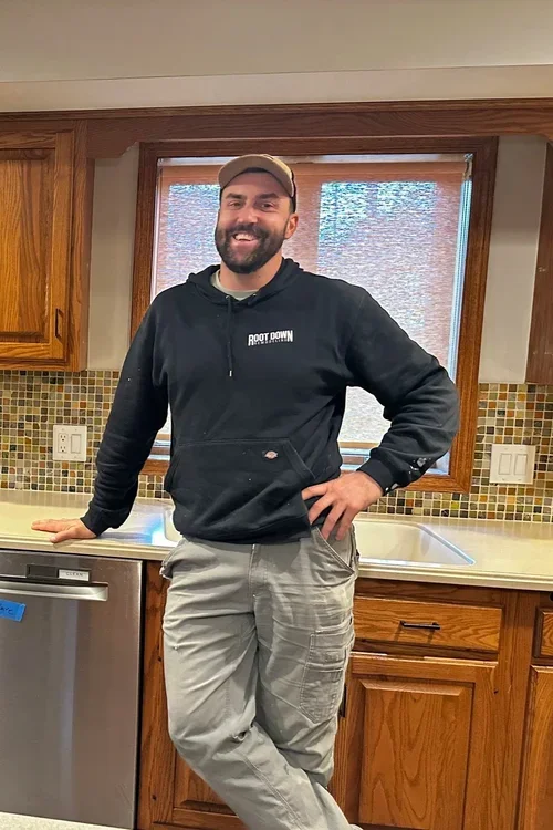 A man with a beard and cap standing in a kitchen, smiling, wearing a black hoodie and gray cargo pants.