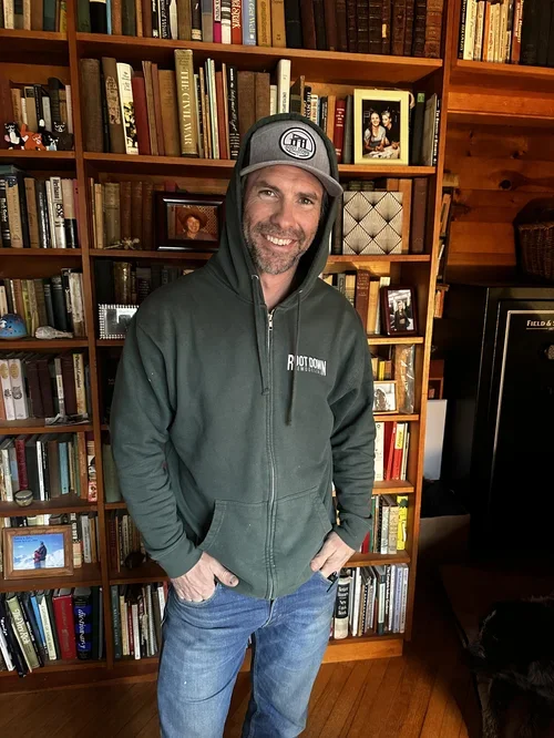 A man standing in front of a wooden bookshelf filled with books, wearing a gray hoodie with a logo, a cap, and jeans, smiling with hands in pockets.