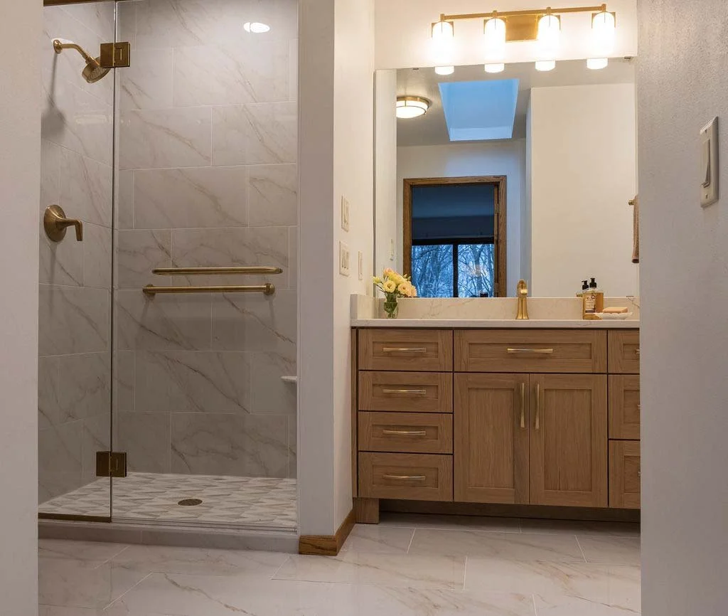 Bathroom with a walk-in shower featuring gold fixtures and a wooden vanity with a large mirror and bright lighting.