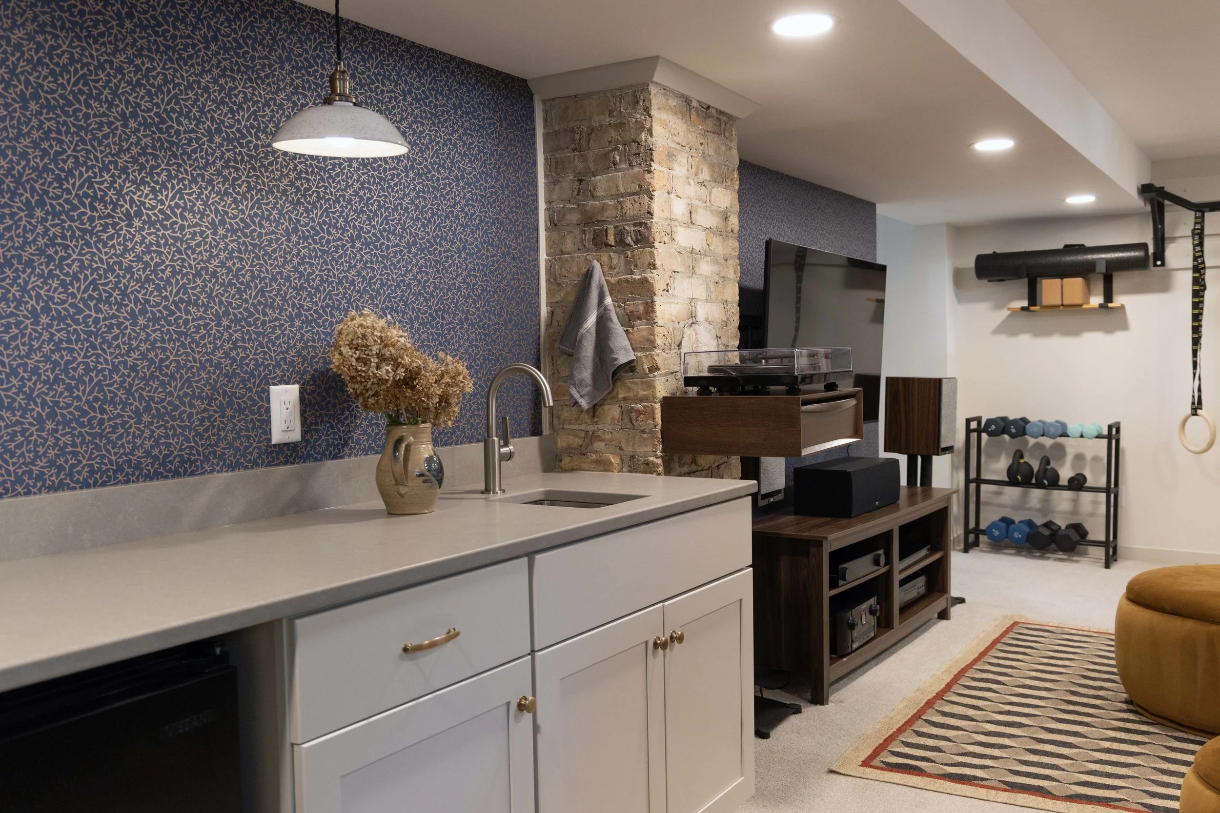 Finished basement wet bar featuring a built-in sink, quartz counters, dark wallpaper, and an exposed brick accent wall.