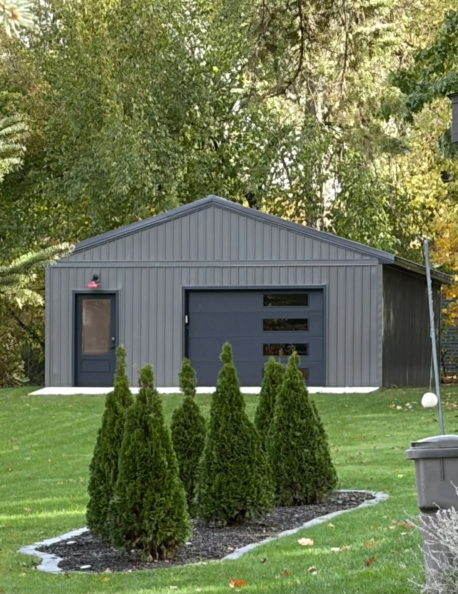 A small gray building with a gabled roof and a garage door, surrounded by a neatly landscaped yard with five evergreen trees planted in a row and a small garden bed. Felled leaves are visible on the grass, suggesting autumn.