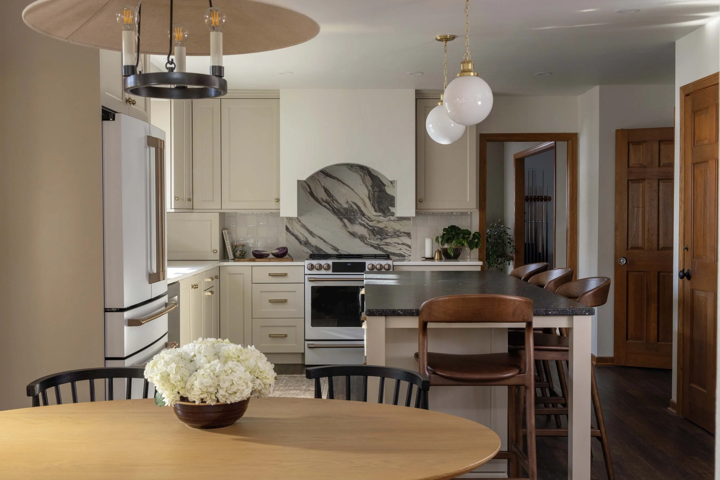 A modern kitchen with cream-colored cabinets, a marble backsplash, and a stainless steel stove. There are two white globe pendant lights hanging from the ceiling, a wooden door, and a black countertop island with brown chairs. In the foreground, there is a table with a vase of white flowers.
