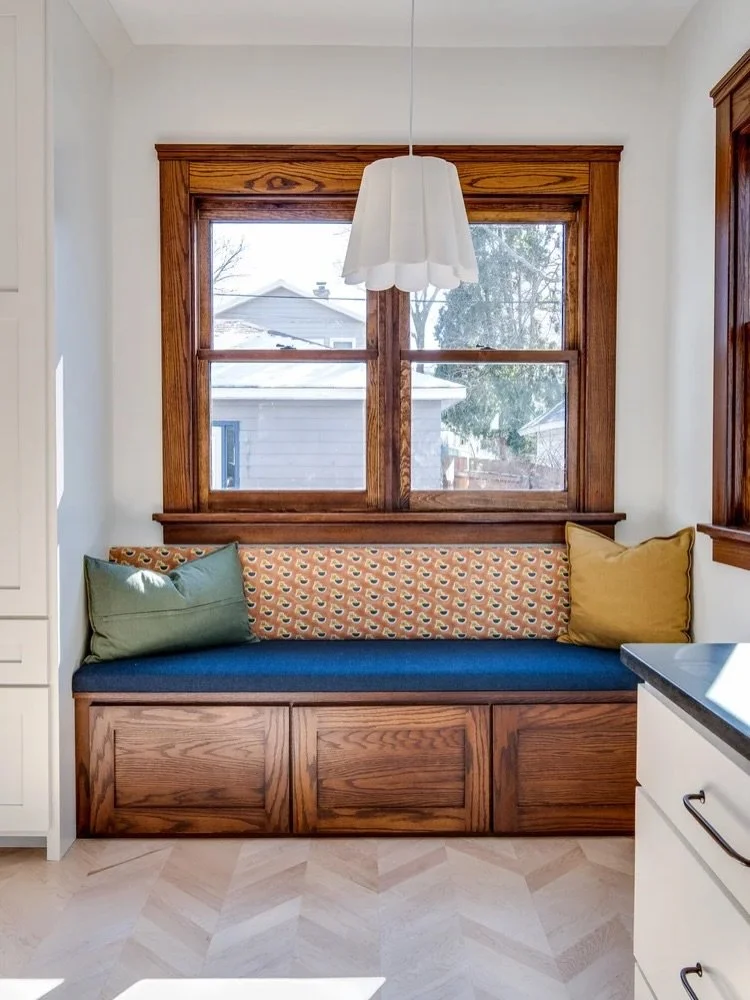 A built-in window seat with colorful cushions and a large window with wooden trim, inside a white kitchen area.