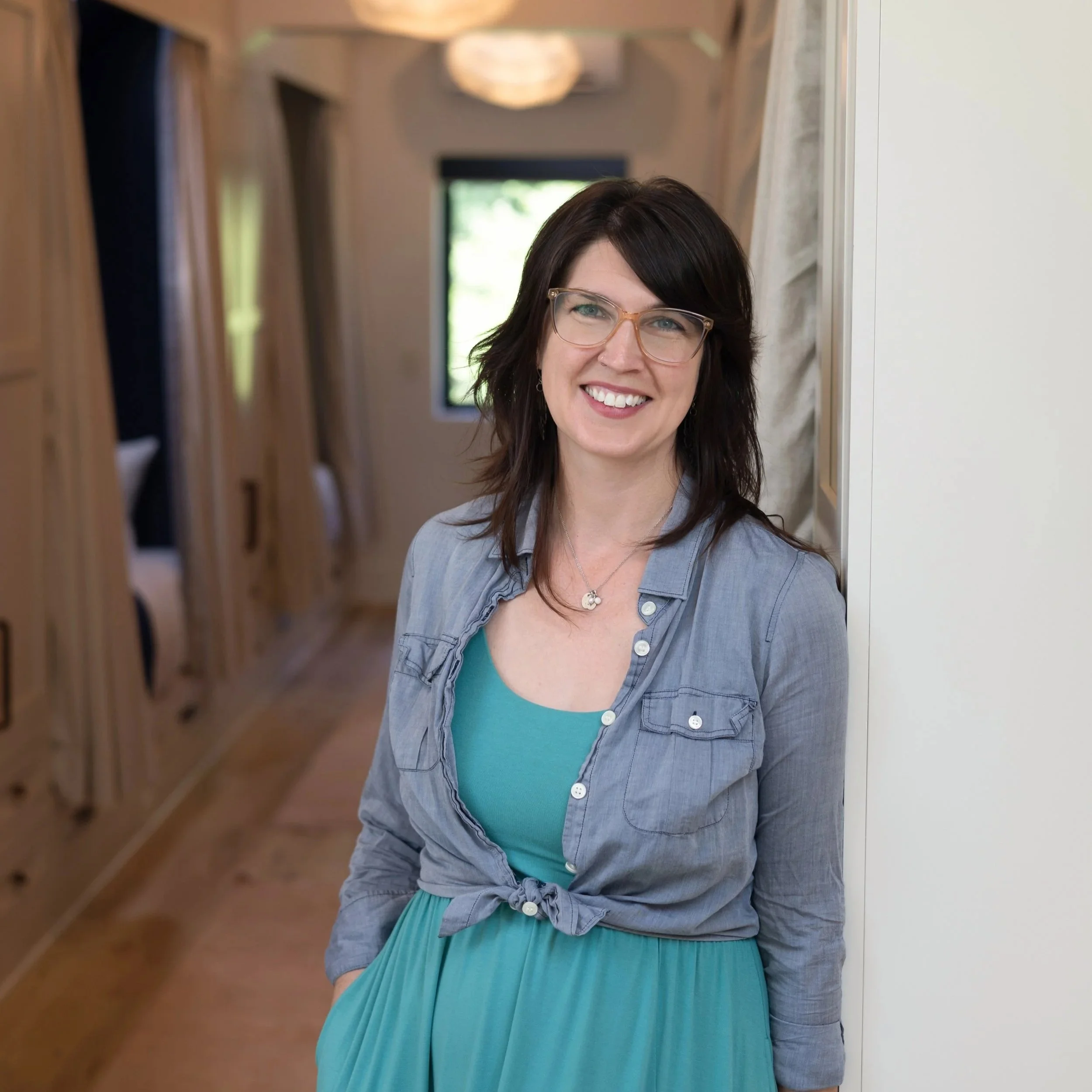 A smiling woman with dark brown hair and glasses, wearing a blue dress and denim shirt, standing in a hallway with wooden shelves on one side and a door on the other.