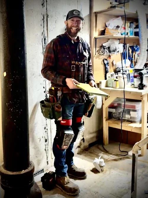 A man with a beard smiling in a workshop, holding a yellow clipboard, wearing a plaid shirt, jeans, work boots, and a tool belt, surrounded by tools and supplies on shelves.