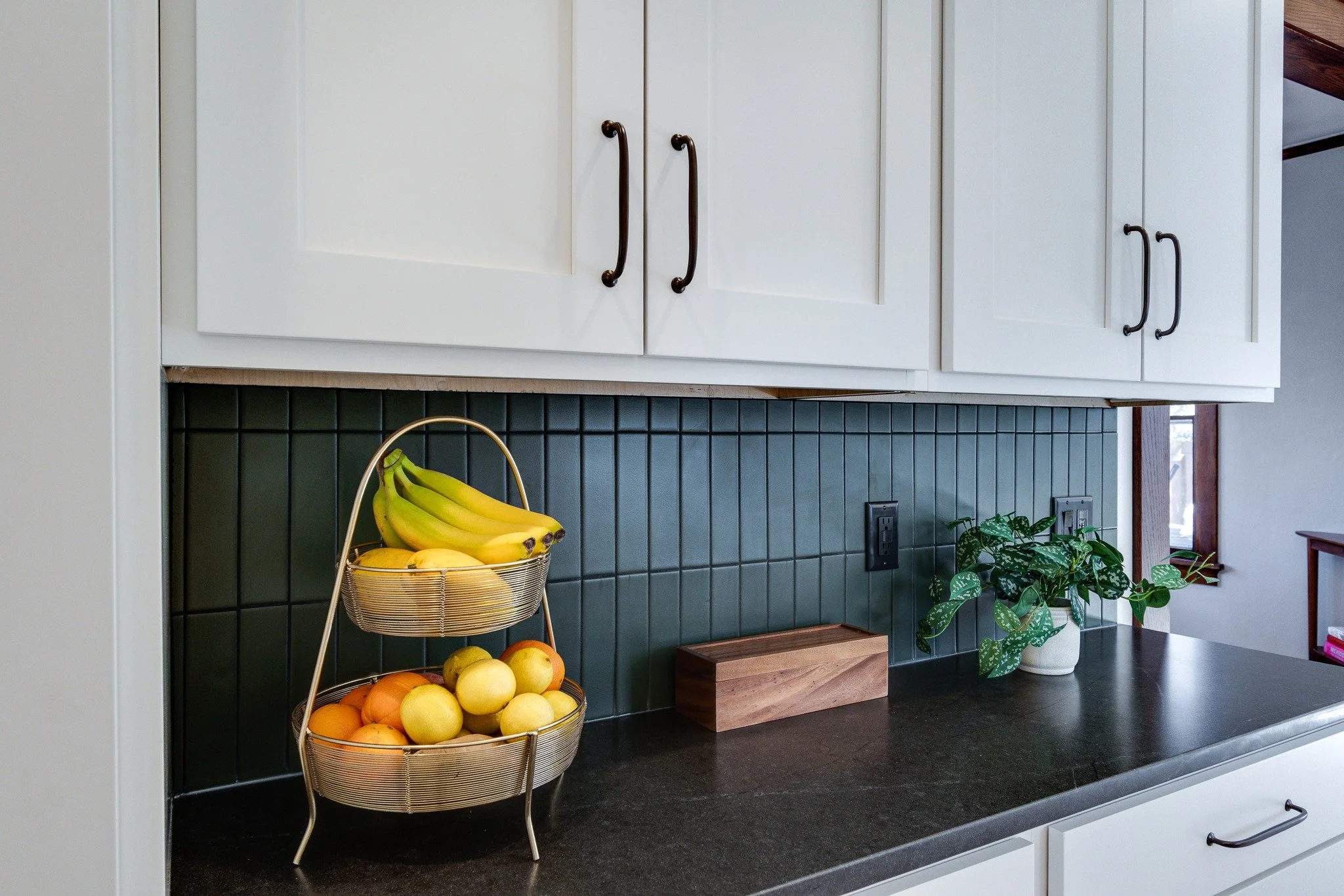 Kitchen with white cabinets, dark countertop, green tiled backsplash, and a basket of bananas and apples on the counter.
