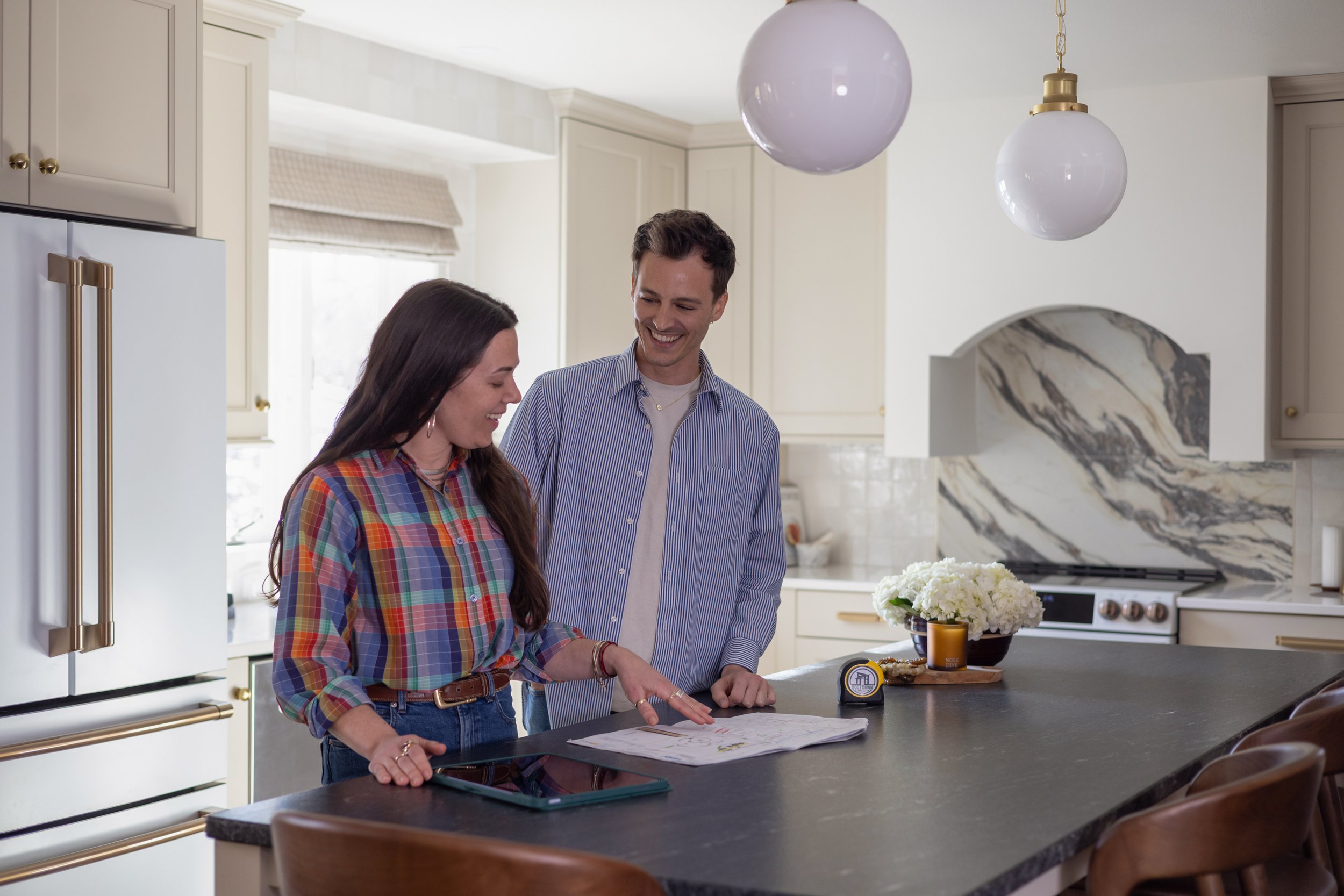 A man and a woman smiling and looking at a paper on a kitchen counter, with a bouquet of white flowers and a small candle nearby.