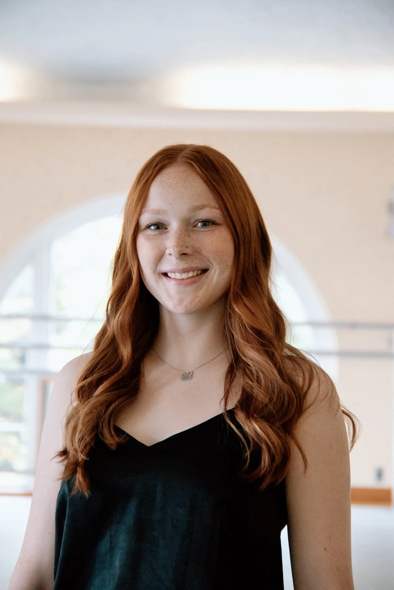 A young woman with long, wavy red hair and fair skin, smiling at the camera. She is wearing a black sleeveless top and a delicate necklace, standing in front of a large arched window in a well-lit room.