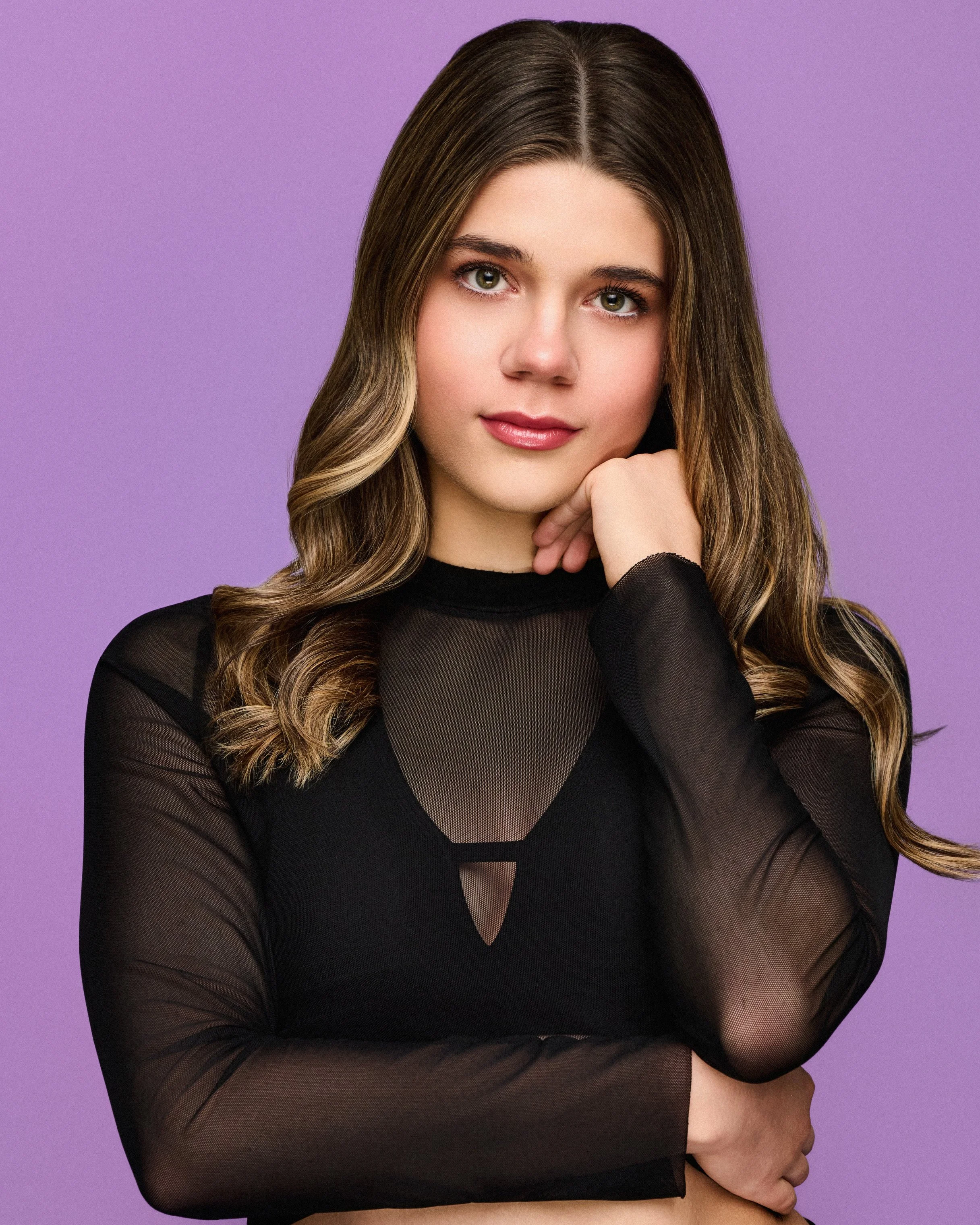 A young woman with long wavy brown hair, wearing a black sheer long-sleeve top, resting her chin on her hand, against a purple background.