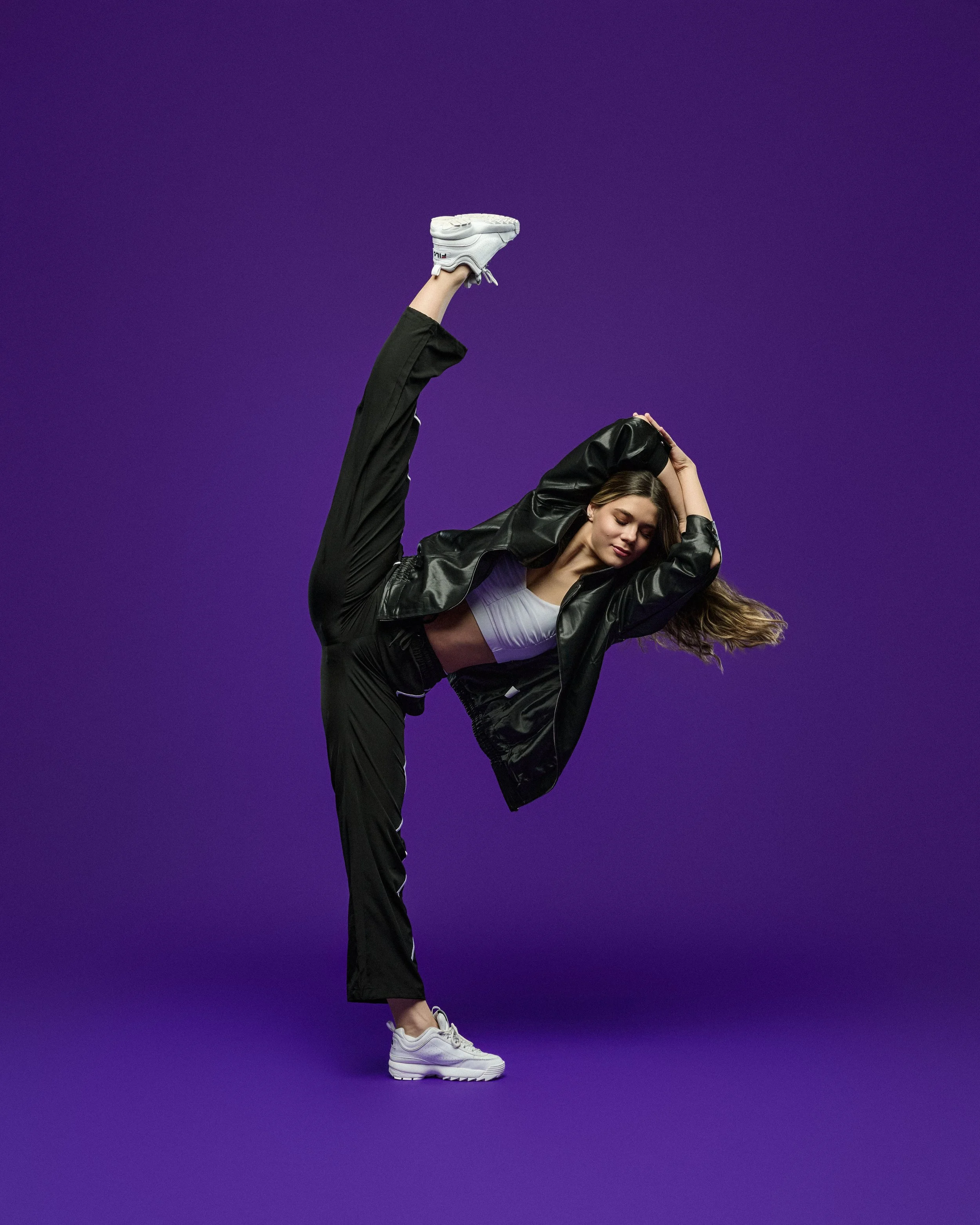 A woman in black jacket and pants stretching on a purple background, balancing on one leg with the other leg raised high, wearing white sneakers, long hair flowing to the side, eyes closed, appearing focused and relaxed.