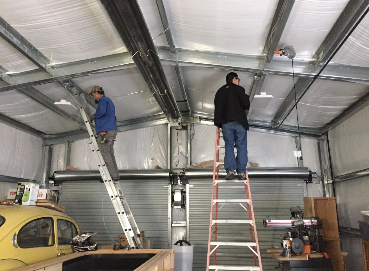 Two men working on the ceiling of an industrial garage or workshop. One standing on an aluminum ladder, the other on an orange ladder. The garage contains various tools and a yellow vehicle.