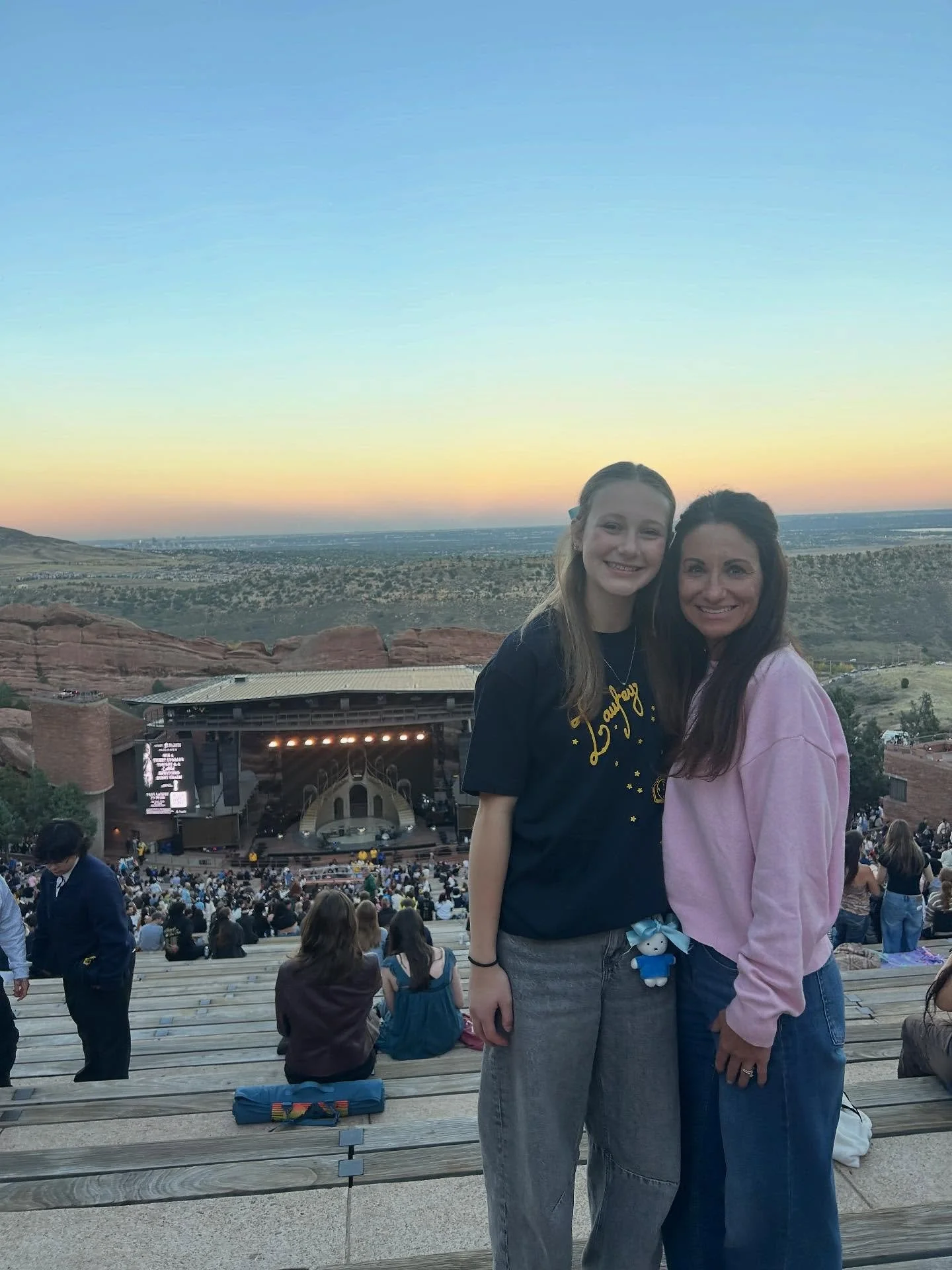 Two women standing close together on an outdoor amphitheater stage with a scenic mountain and sunset background, smiling at the camera, surrounded by a seated audience.