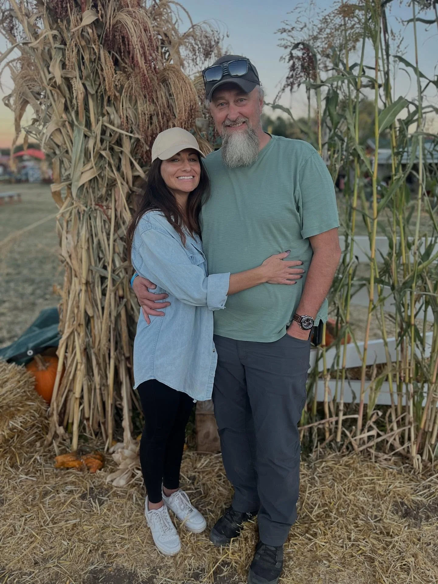 A smiling couple standing outdoors at a pumpkin patch, surrounded by dried cornstalks and pumpkins, with trees and a clear sky in the background.