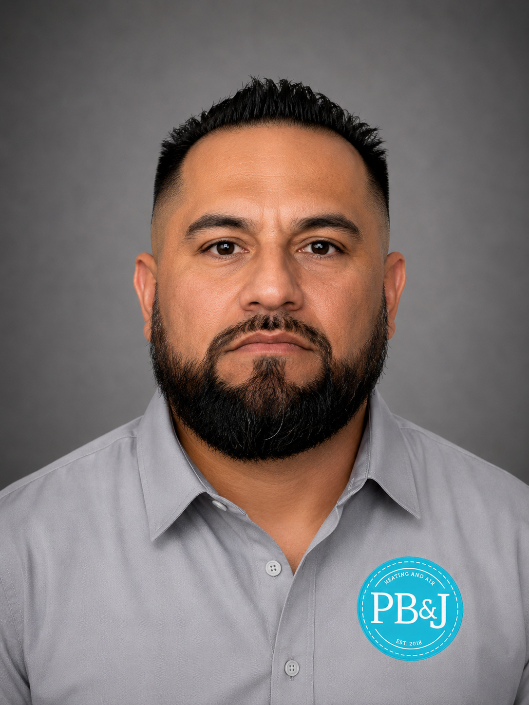 A man with a dark beard and mustache, wearing a light gray collared shirt with a blue logo on the chest that reads 'PB&J Heating and Air, Est. 2018,' against a gray background.
