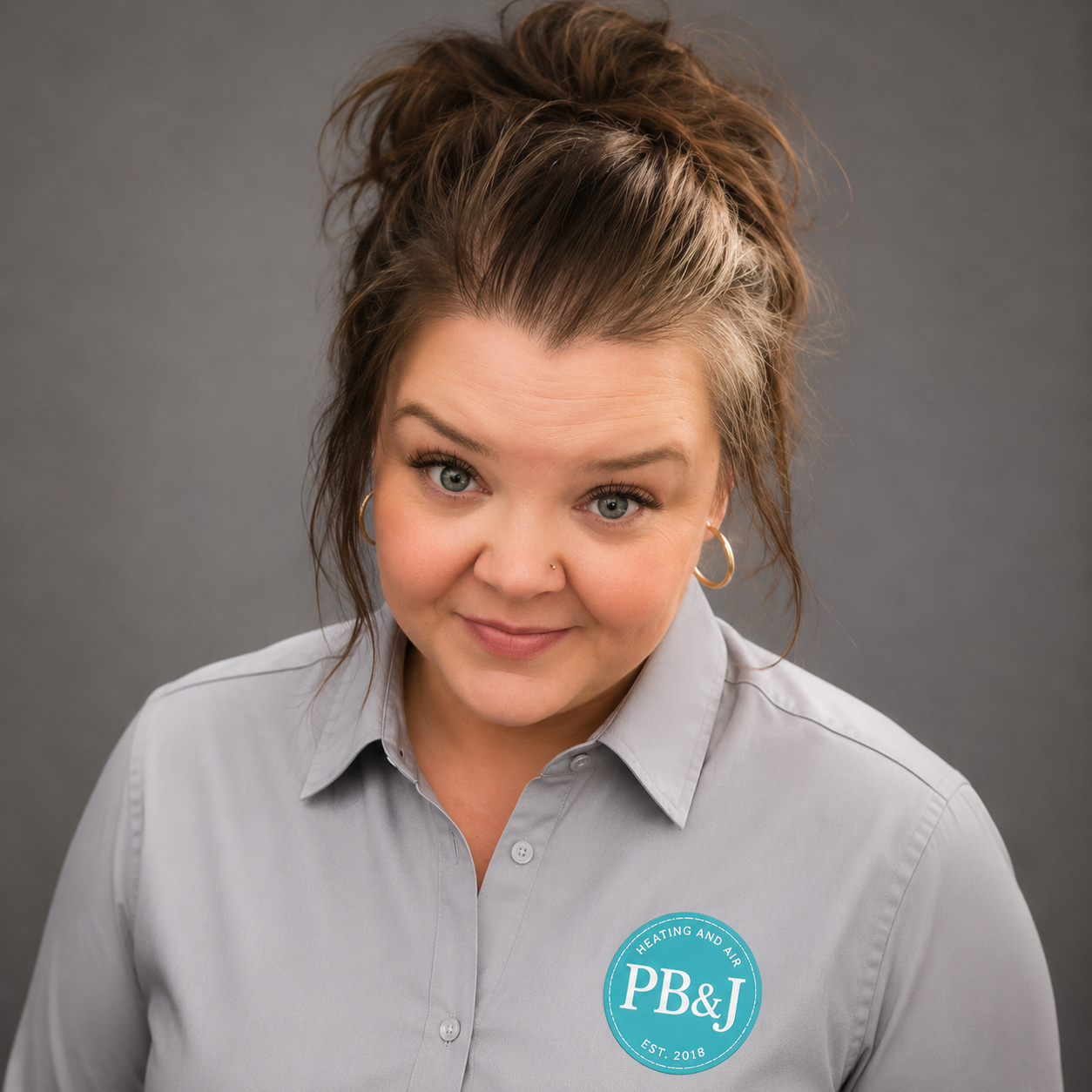 A woman with curly brown hair in a bun, wearing a light gray button-up shirt with a blue logo reading "PB&J Heating and Air, Est. 2018" on the shirt, looking at the camera with a slight smile.