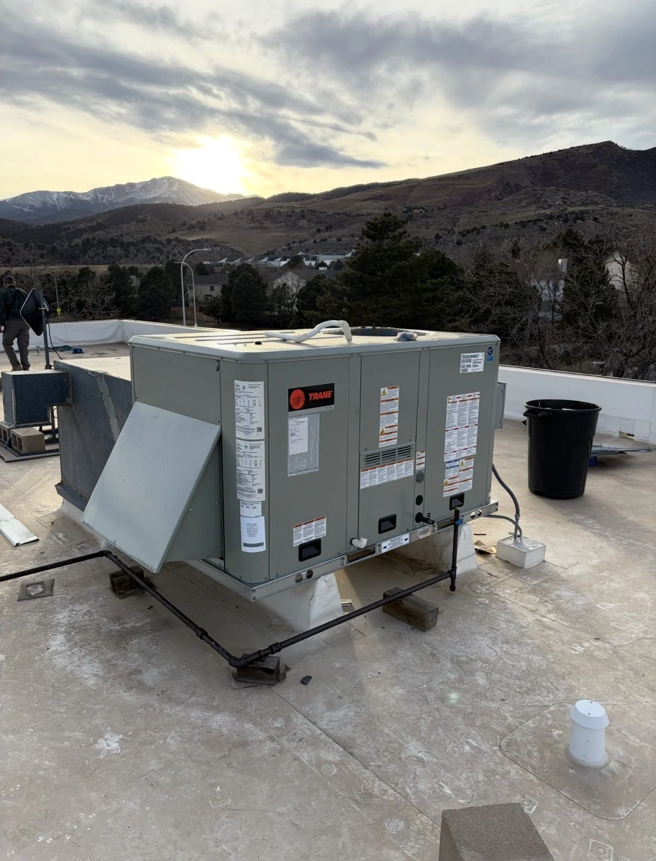 A rooftop with heating, ventilation, and air conditioning (HVAC) equipment, including a Trane air handler, with a scenic mountain landscape in the background during sunset.