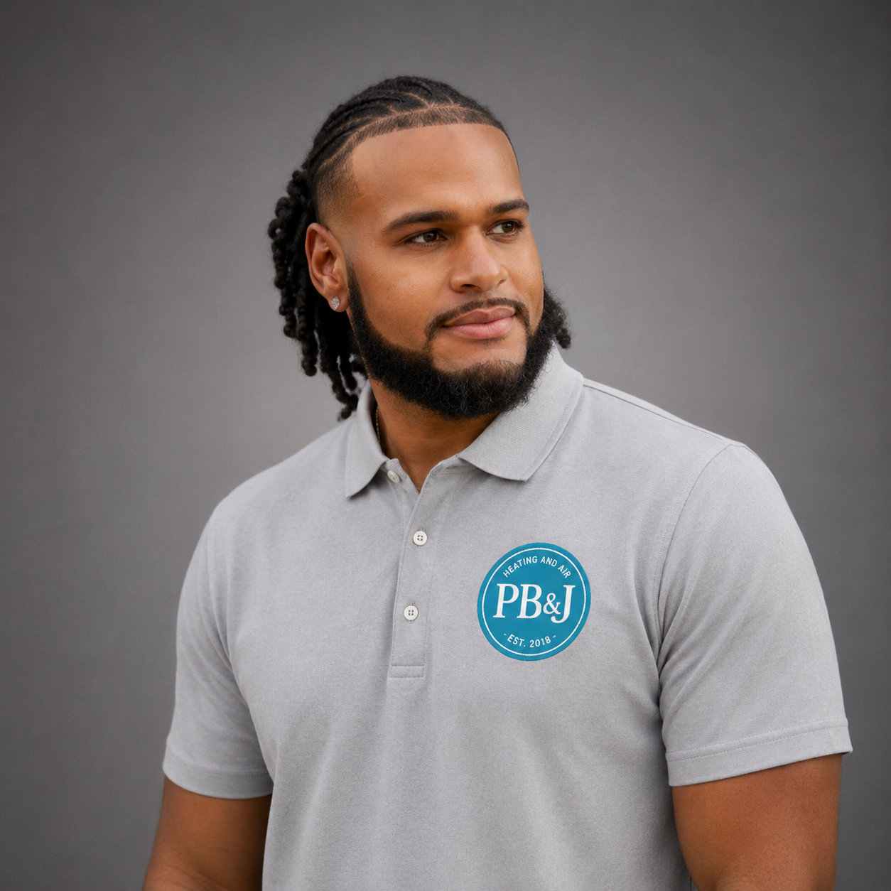 A young man with a beard and dreadlocks wearing a light gray polo shirt with a blue logo that reads 'PB&J Heating and Air' established in 2018, looking to the side against a gray background.