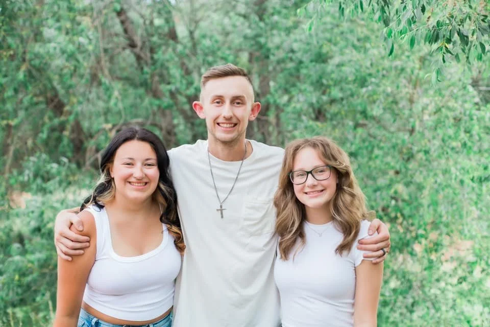 Three smiling people standing outdoors with their arms around each other, surrounded by green trees.