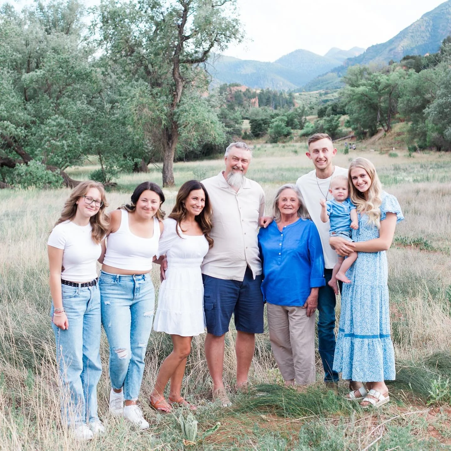 A family of nine people standing outdoors in a grassy field with trees and mountains in the background, smiling at the camera.