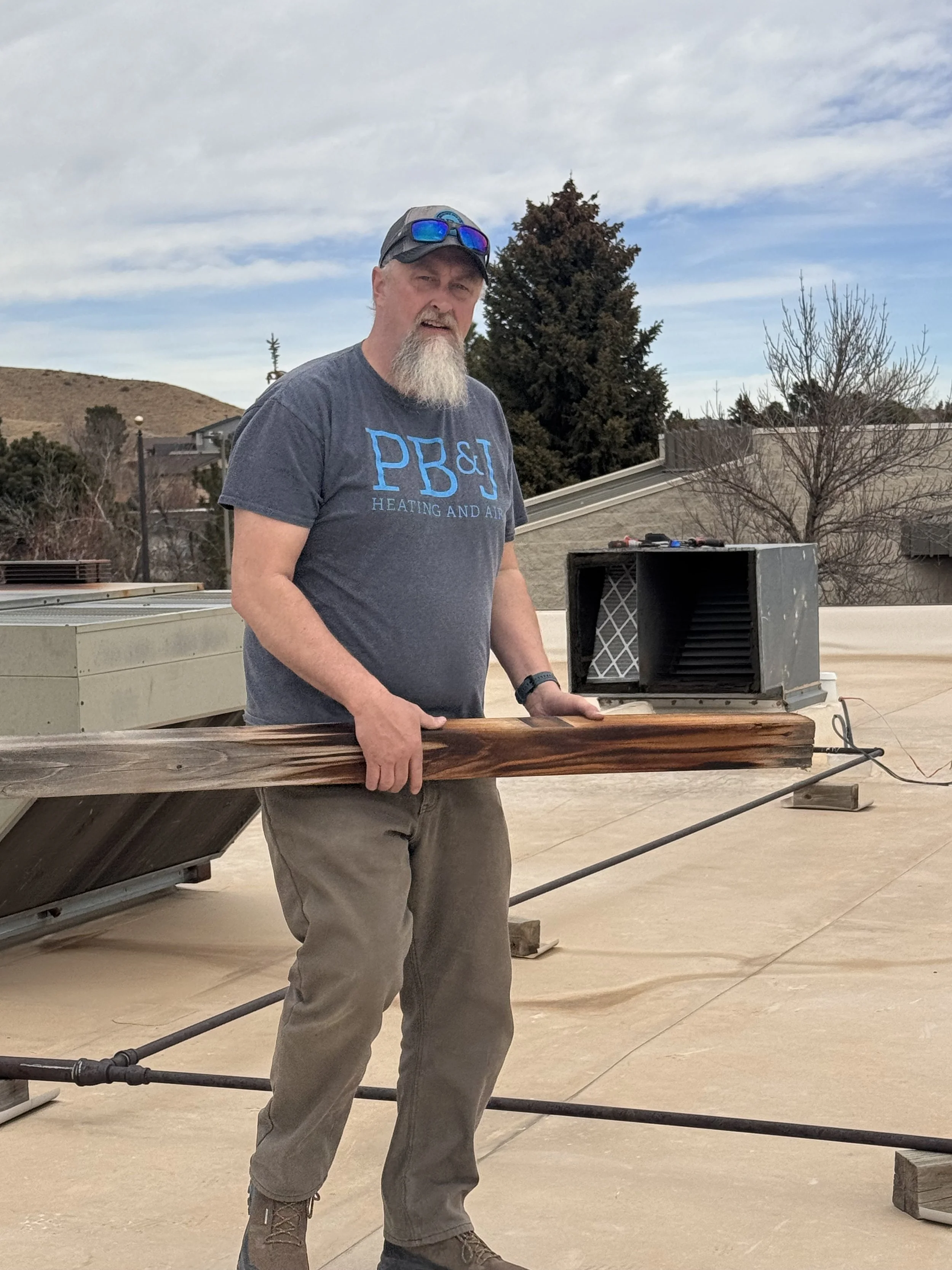 A man with a gray beard and wearing a black cap, sunglasses, a dark gray T-shirt with blue letters, tan pants, and boots, standing outdoors on a rooftop with a speaker and a piece of wood in front of him, with trees and buildings in the background.