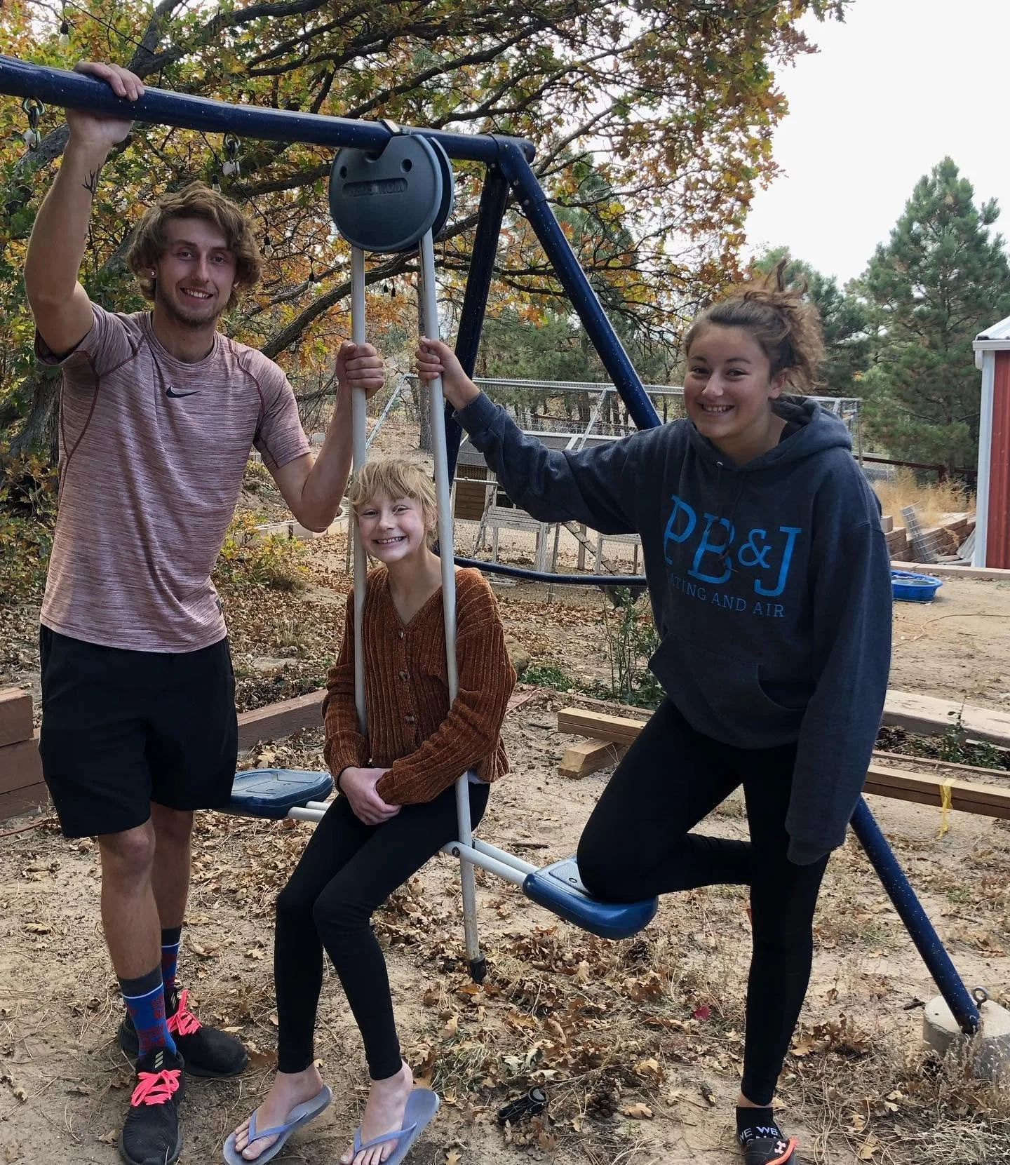 Four people standing outdoors next to a swing set, smiling at the camera. The background has trees with autumn leaves, and a red shed is visible to the right.