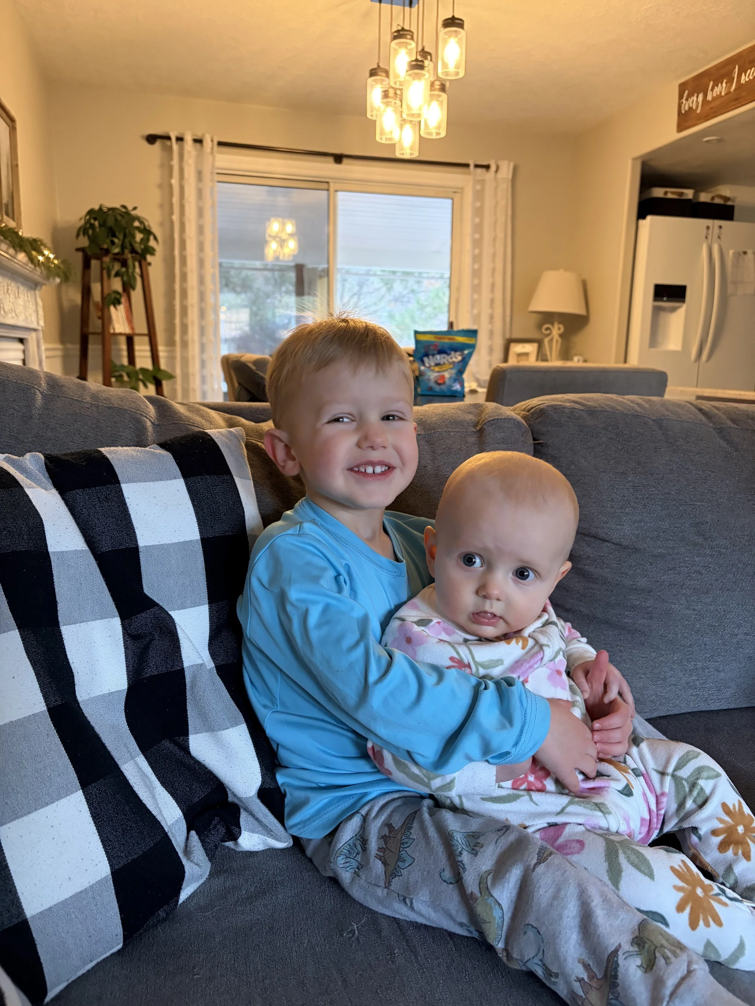 Two young children sitting on a gray sofa, smiling at the camera. A boy with light brown hair in a blue shirt is hugging a baby with big blue eyes, dressed in floral pajamas. The background shows a living room with a window, a contemporary chandelier, and holiday decorations.