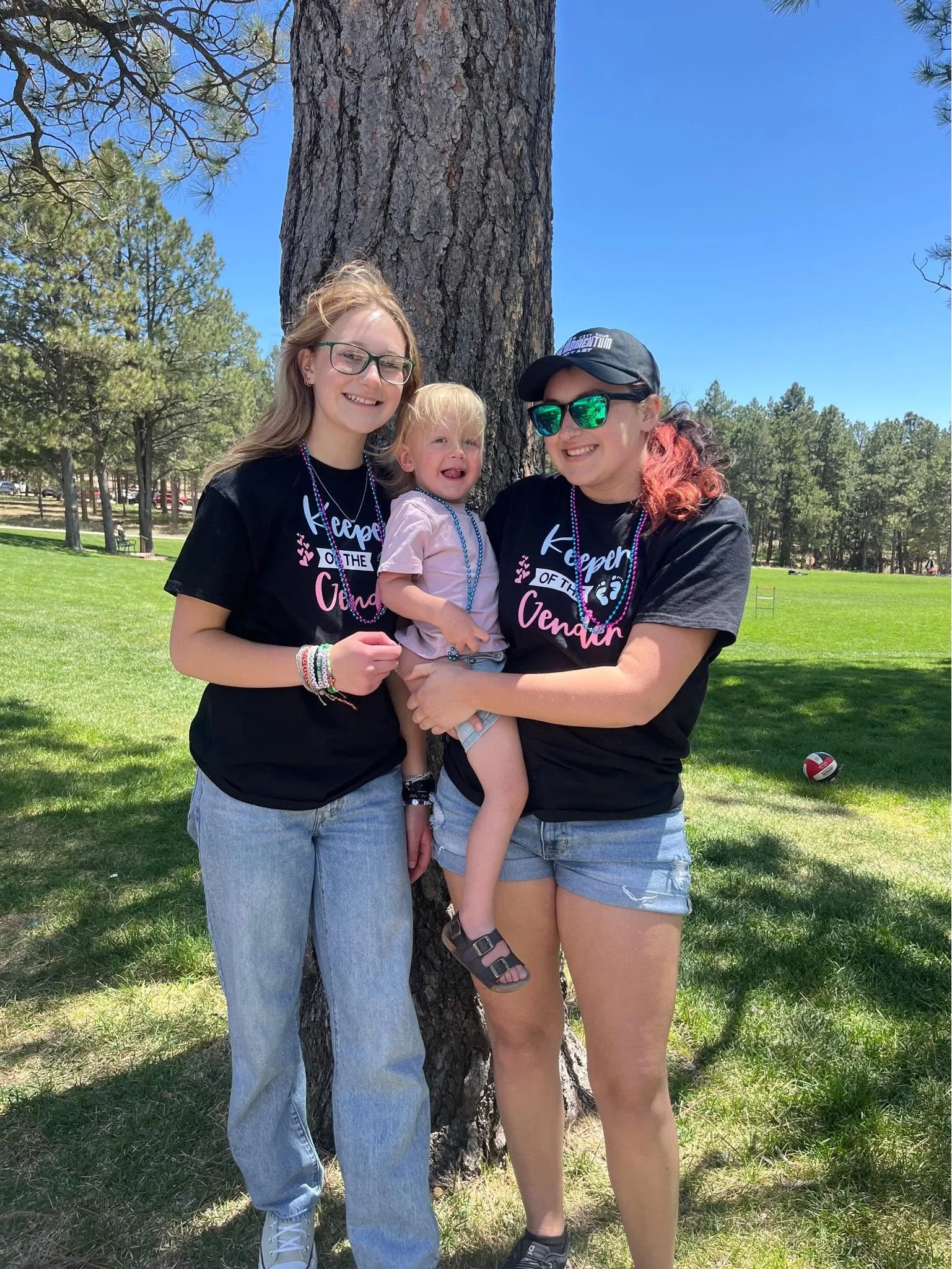 Three women and a child standing outdoors in front of a large tree with a park in the background under a clear blue sky. Two women are wearing black T-shirts with pink and white text, one woman has red hair and sunglasses, and the other has blonde hair and glasses. The woman with red hair is holding a young girl with blonde hair, while the other woman stands beside them. All are smiling.