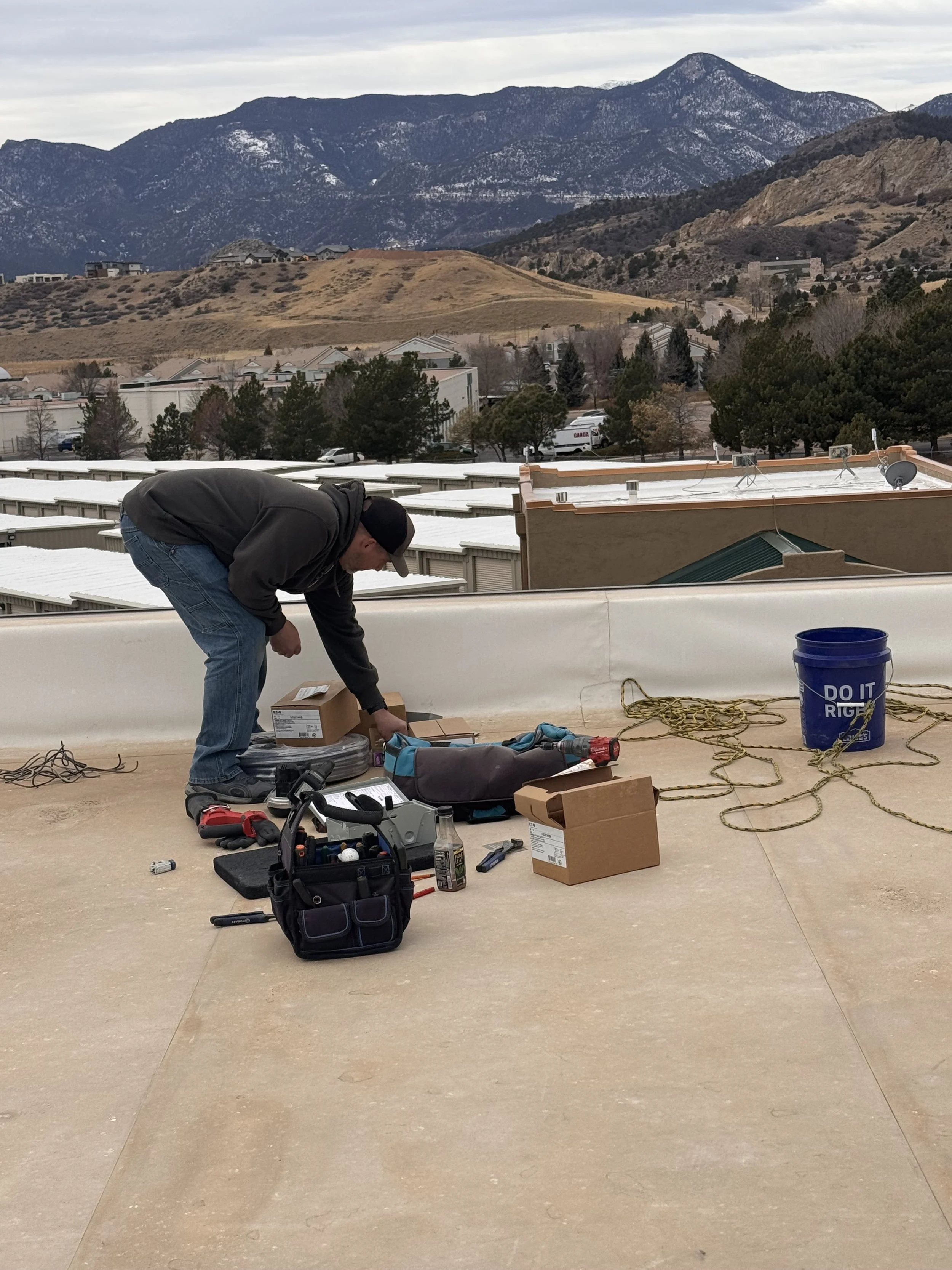 A person crouching on a rooftop with various tools and equipment around them, including a toolbox, extension cords, and a bucket. In the background, there are residential buildings, trees, rolling hills, and mountains under a cloudy sky.