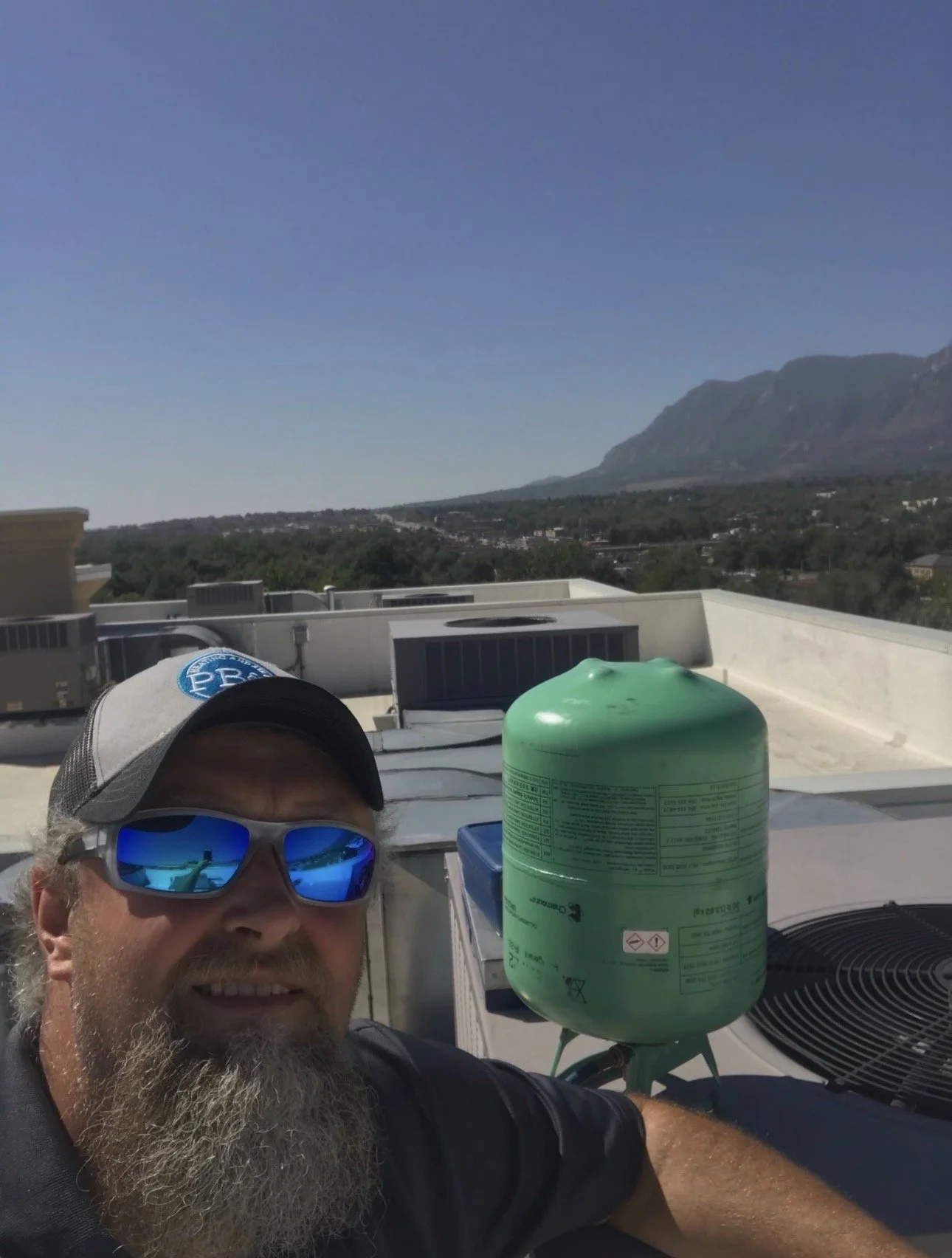A man with a beard wearing sunglasses and a baseball cap taking a selfie on a rooftop. In the background, there are mountain views under a clear blue sky and rooftop equipment including a green propane tank.