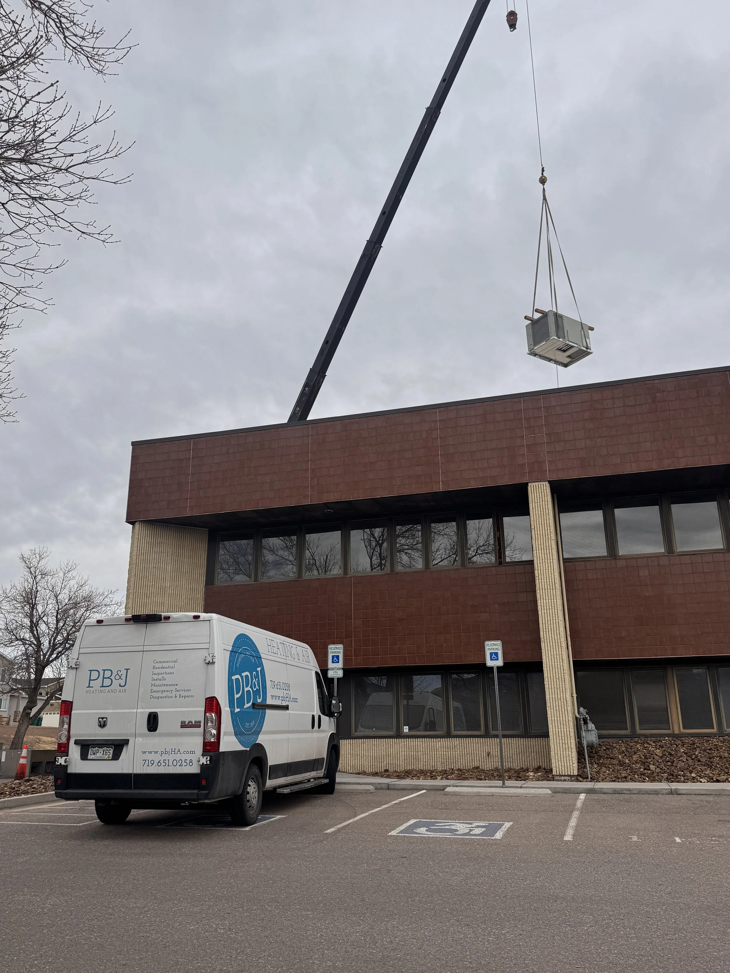 A company van parked in a parking lot with handicapped parking spaces in front of a building. A crane lifts a large HVAC unit to the building's roof.