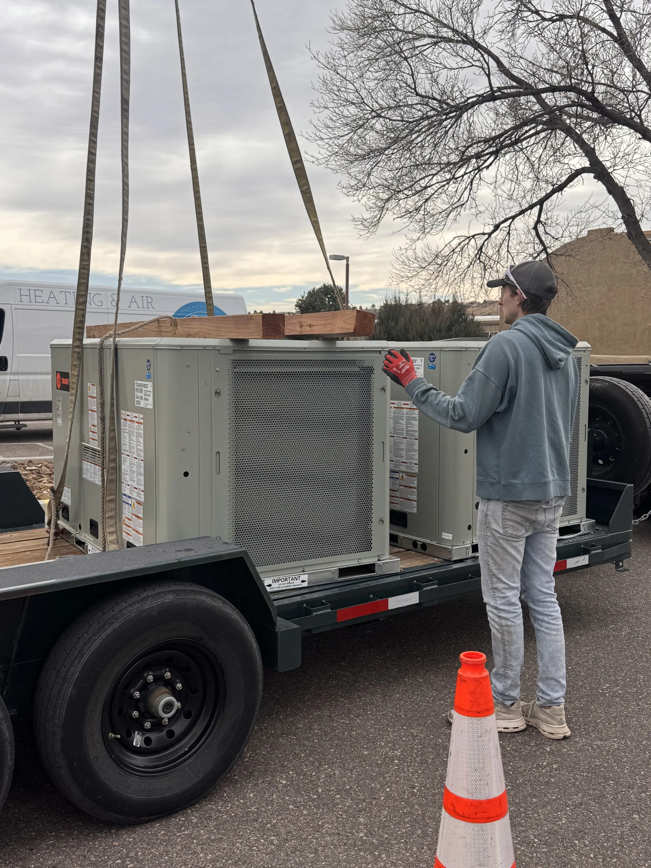 A young man wearing a hoodie, jeans, sunglasses, and red gloves standing on a trailer near a large HVAC unit. He is looking at the equipment while an orange traffic cone is in the foreground. There are leafless trees and a building in the background.