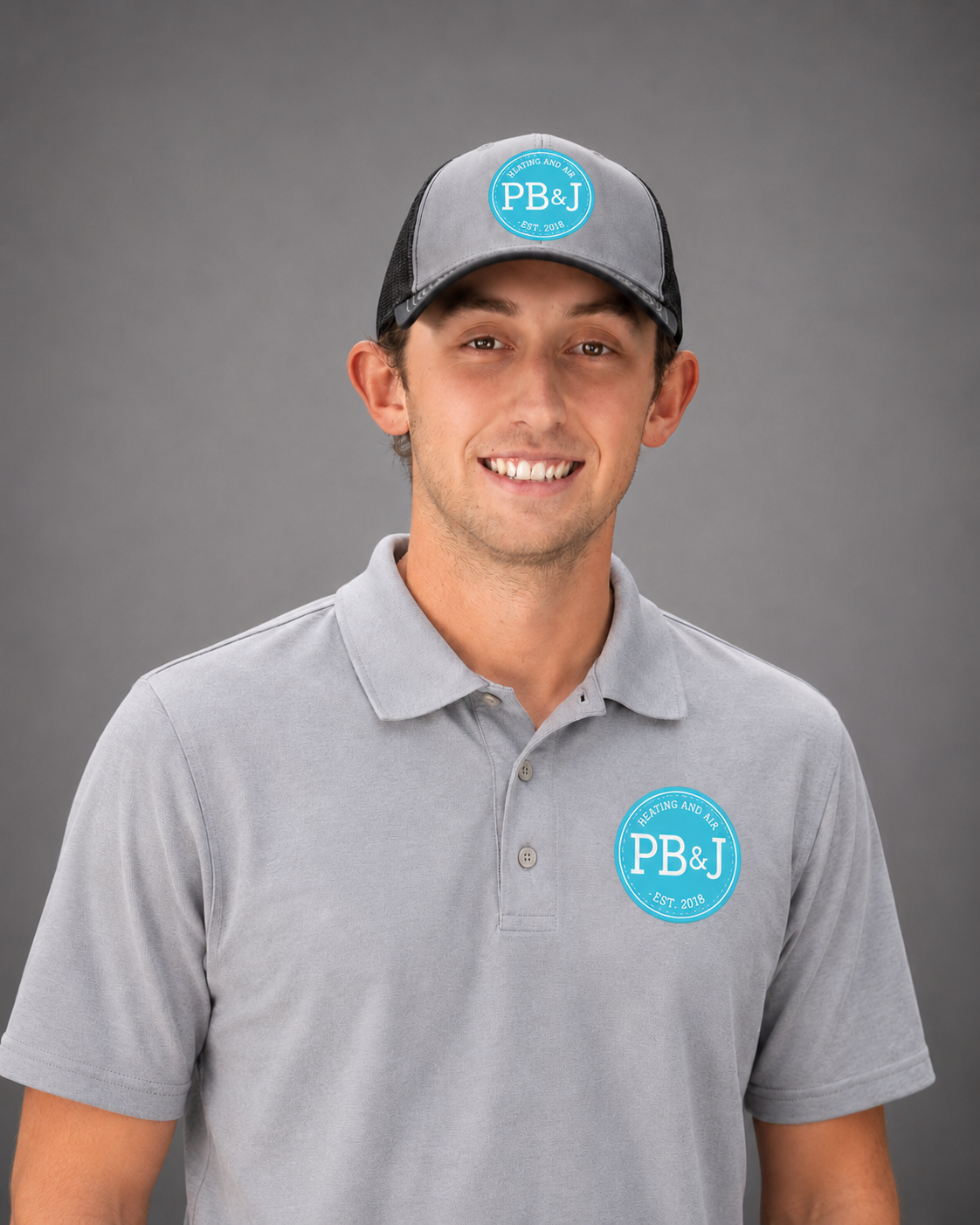 Young man smiling, wearing a grey polo shirt and a grey trucker cap with the logo PB&J, standing against a grey background.