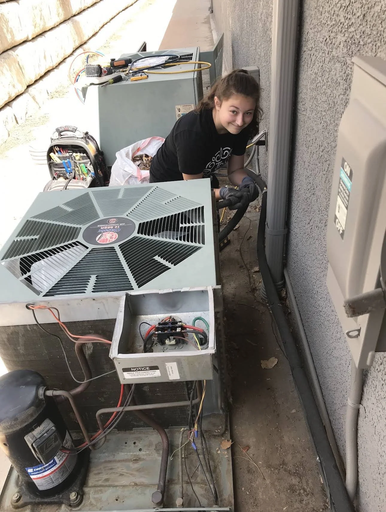 A young woman working on HVAC units outside next to a wall, with tools and equipment around her.