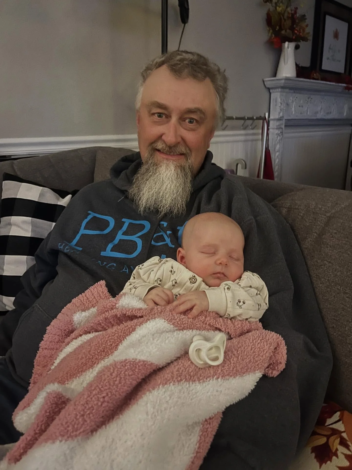 An older man with a beard smiling and holding a sleeping baby wrapped in a pink and white blanket on a couch in a living room.