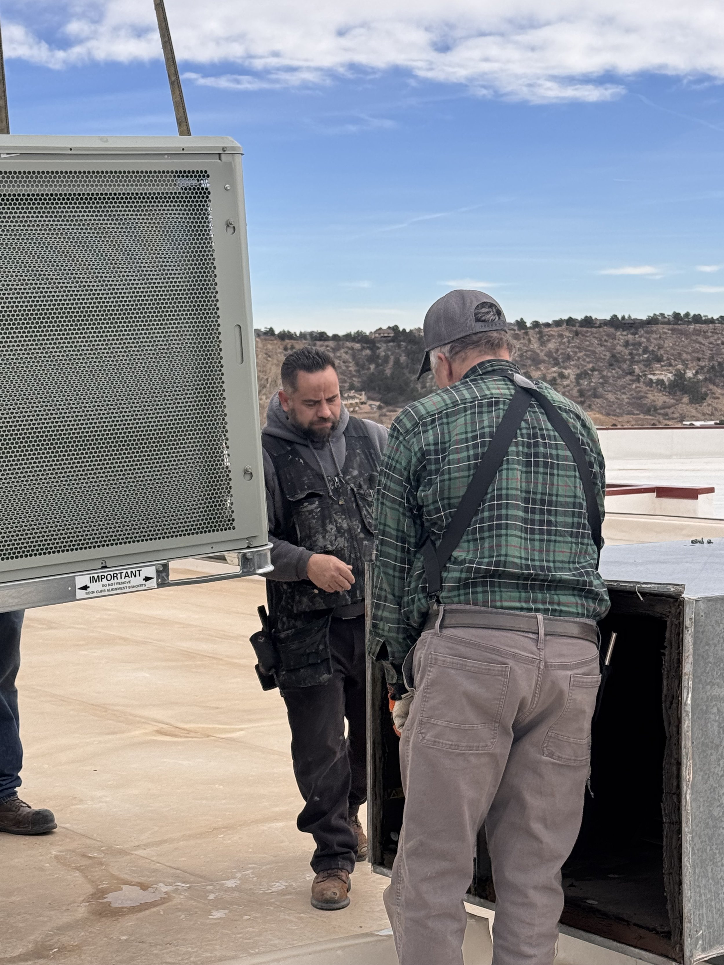 Two men work outdoors near a truck, with a scenic mountainous background, weathered sky, and equipment on a concrete surface.