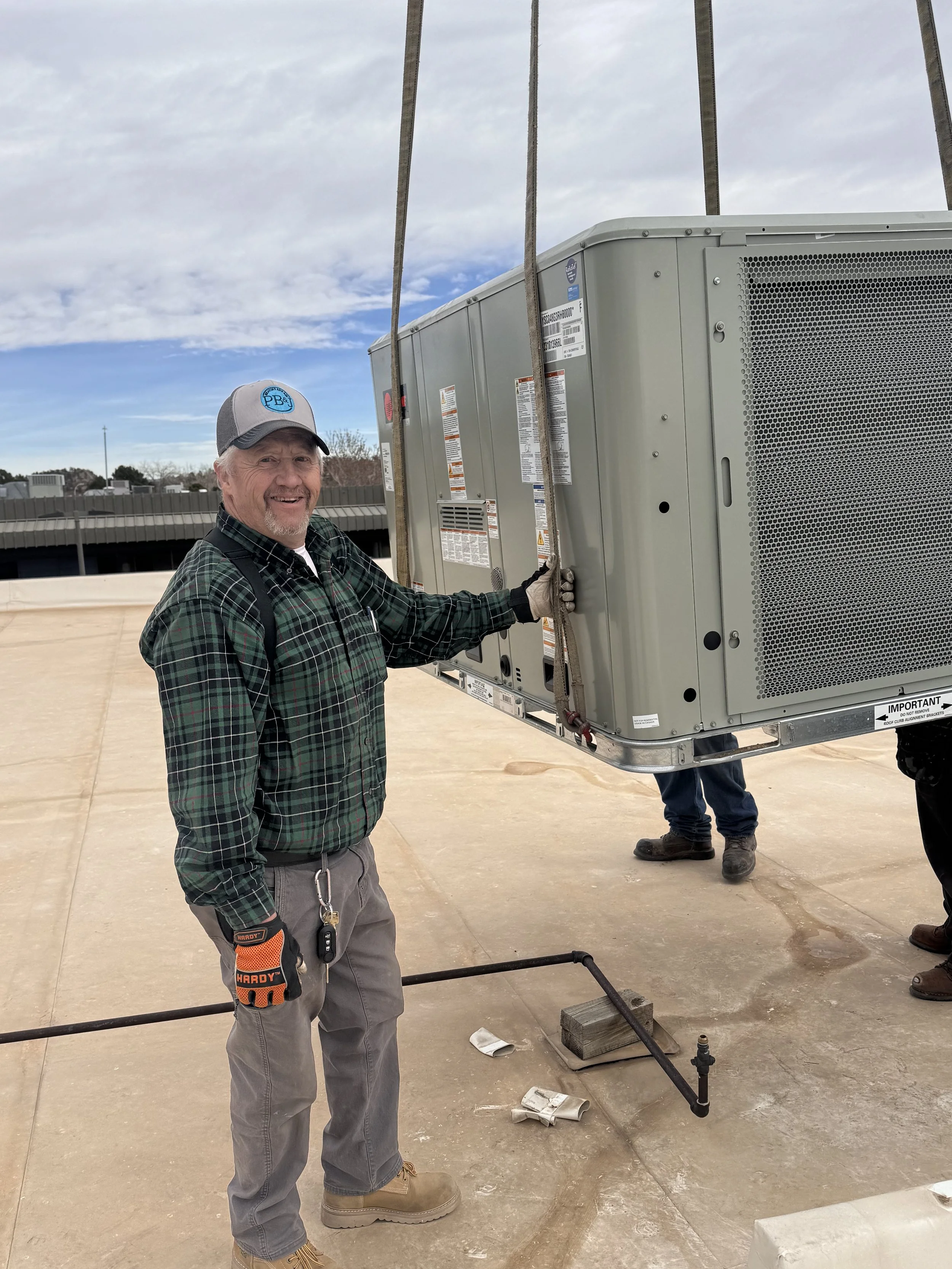 A man in a plaid shirt, gray pants, and work boots stands outdoors near HVAC equipment, smiling at the camera, with a cloudy sky above.