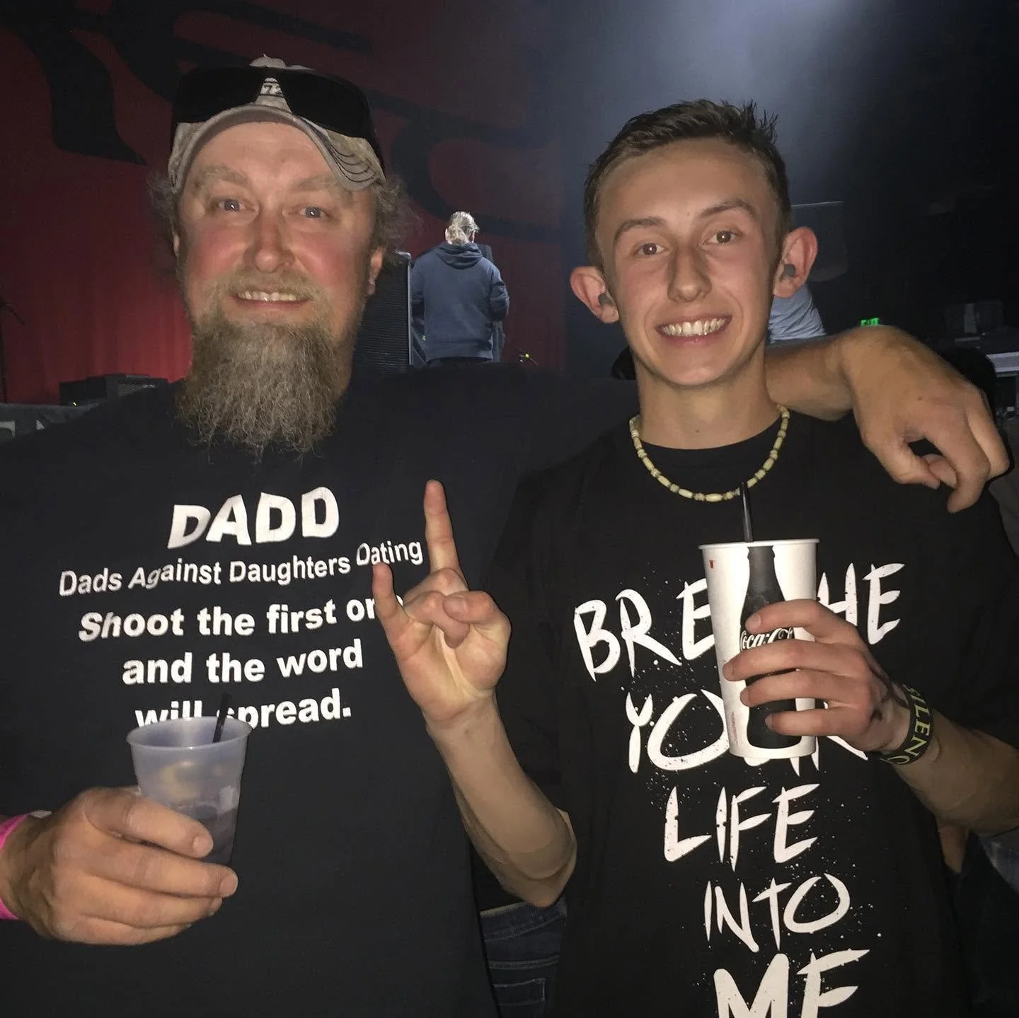 Two smiling males at a party or concert, holding drinks. The man on the left has a beard, glasses on his head, and is wearing a black shirt with the text 'DADS Against Daughters Dating Shoot the first one and the word will spread.' The young man on the right has short hair, a necklace, and is wearing a black shirt with white text, holding a cup with a straw.