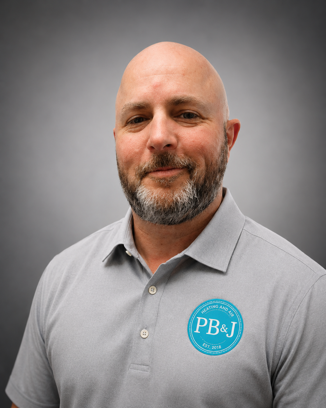 A bald man with a beard wearing a gray polo shirt with a blue circular logo that reads 'PB&J Heating and Air Est. 2018' standing against a plain gray background.