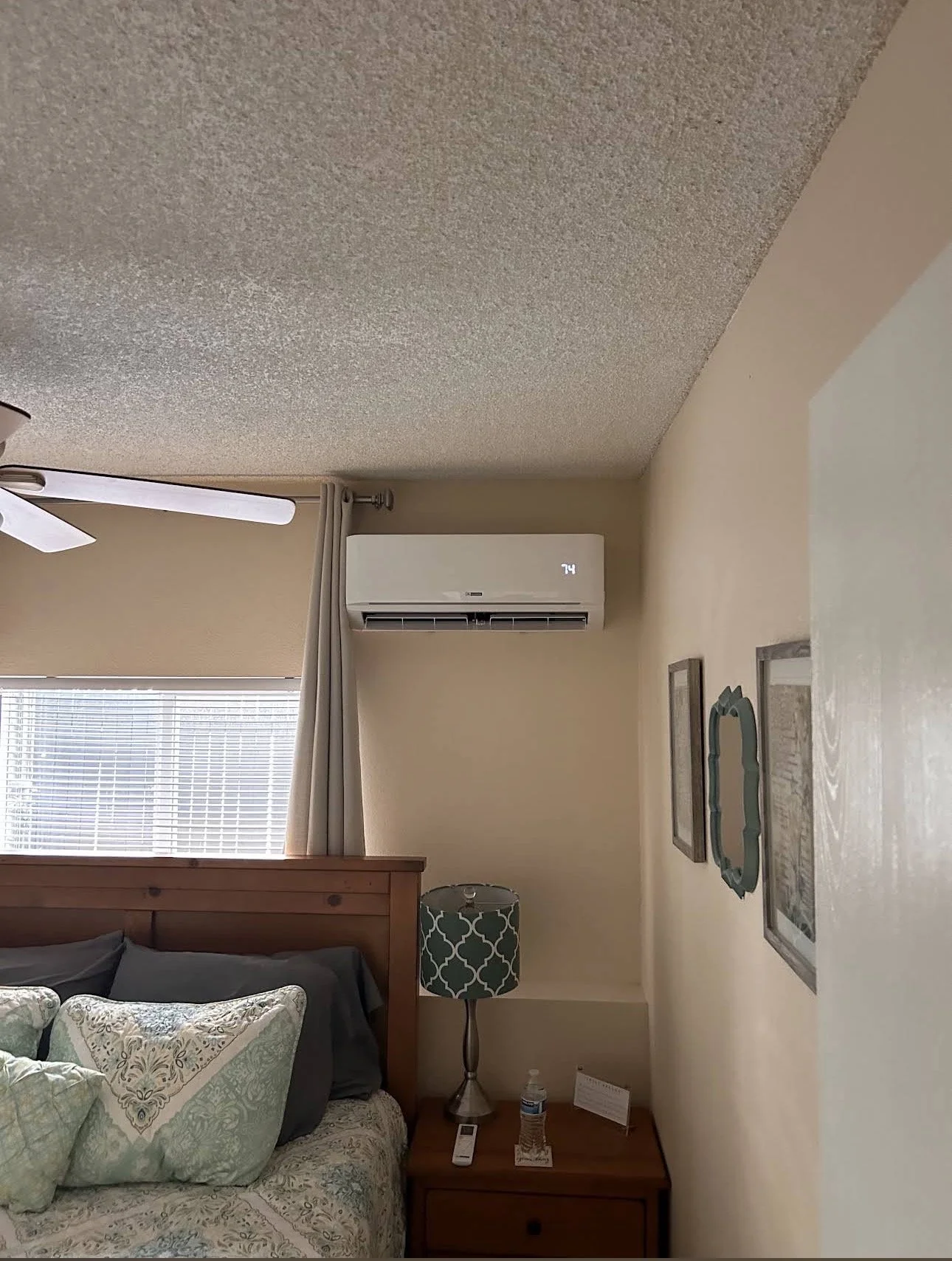 Bedroom corner with a bed, pillows, window with blinds and curtains, an air conditioning unit on the wall, wall art, a nightstand with a lamp, water bottle, TV remote, and a book.
