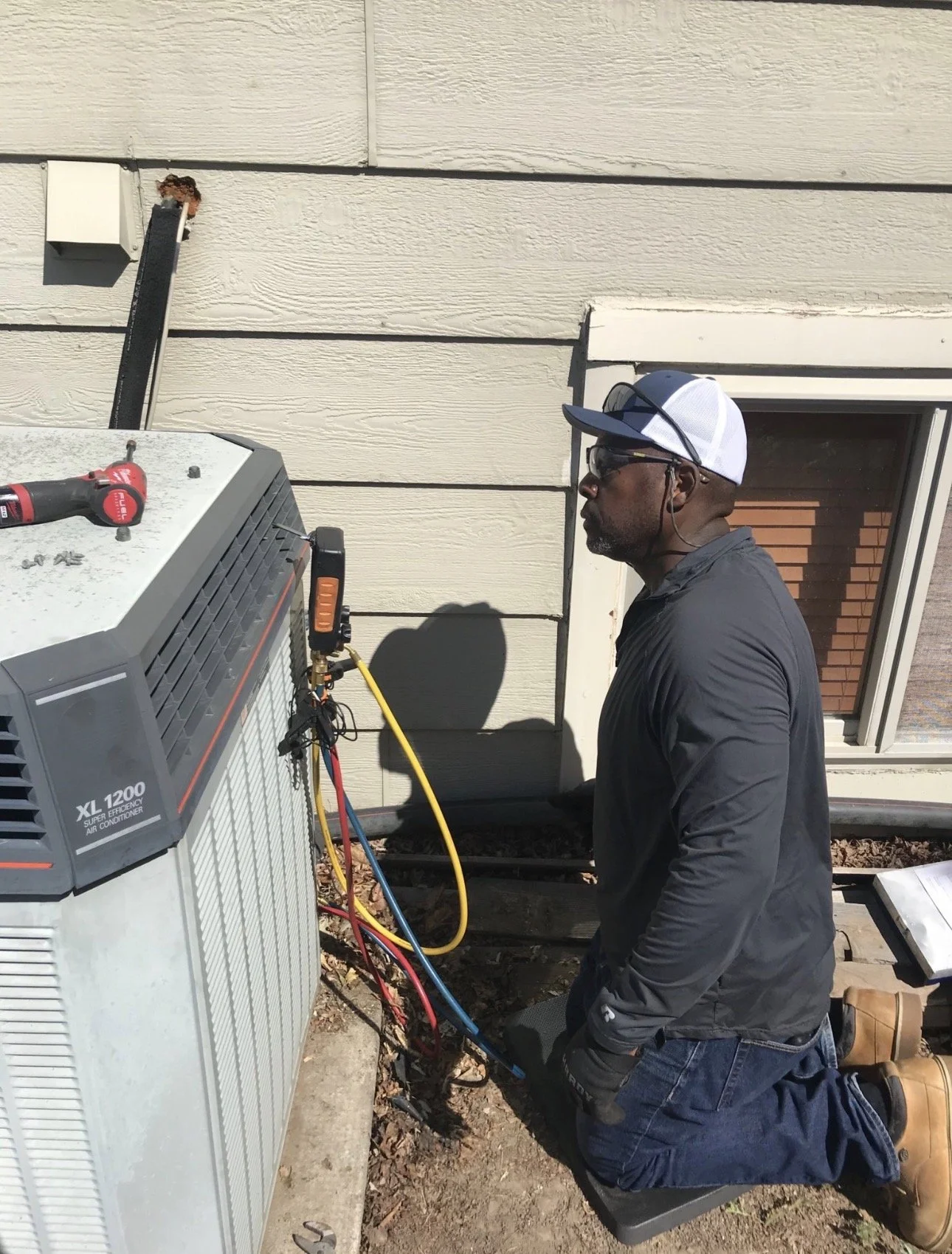 Technician kneeling on a pad working on an outdoor air conditioning unit beside a house wall. He wears safety glasses, a cap, gloves, and a dark jacket. Tools and colored hoses are connected to the unit.