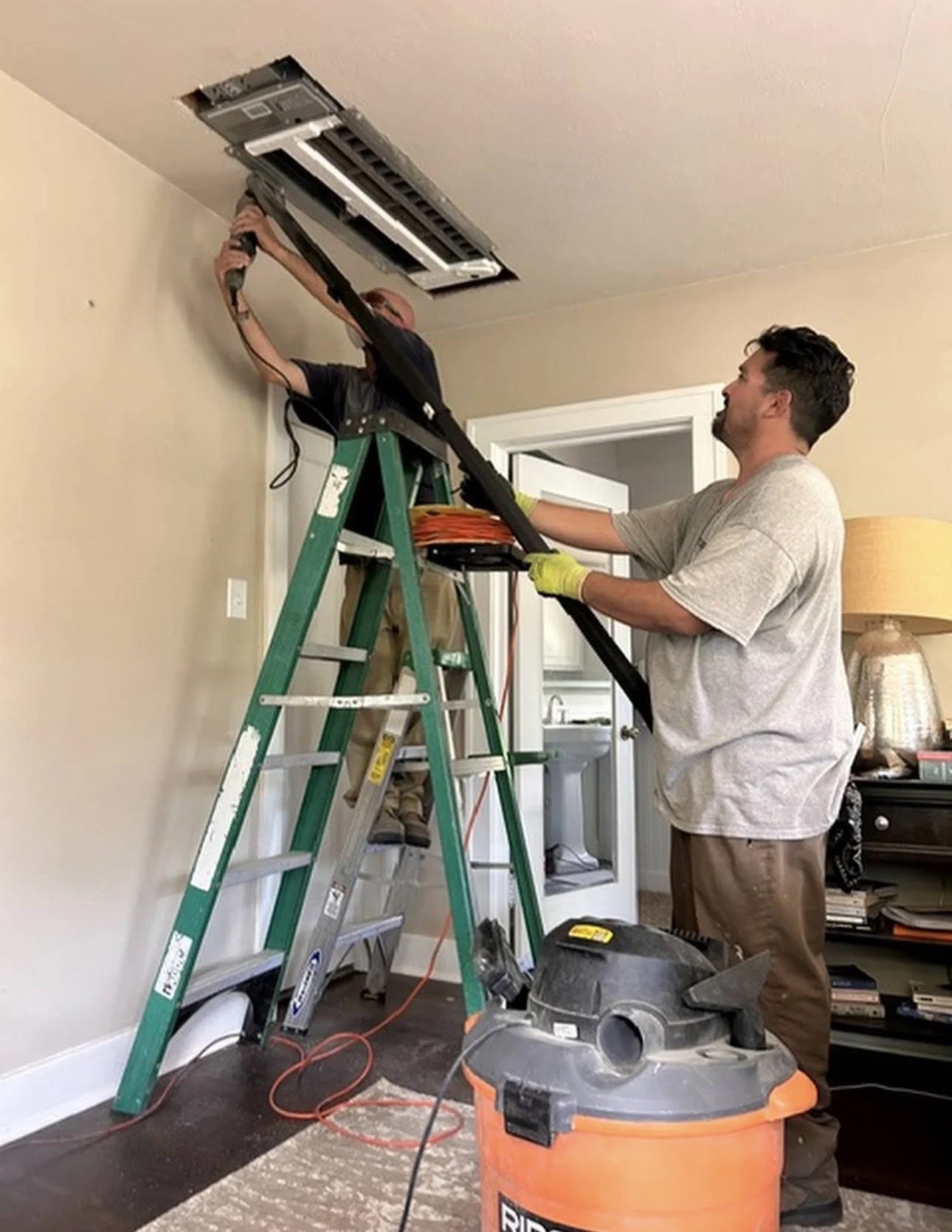 Two men are installing or repairing a ceiling air conditioning vent, one standing on a ladder using tools, the other assisting from the ground. There are tools, extension cords, and a vacuum cleaner nearby in a room with an open doorway and furniture